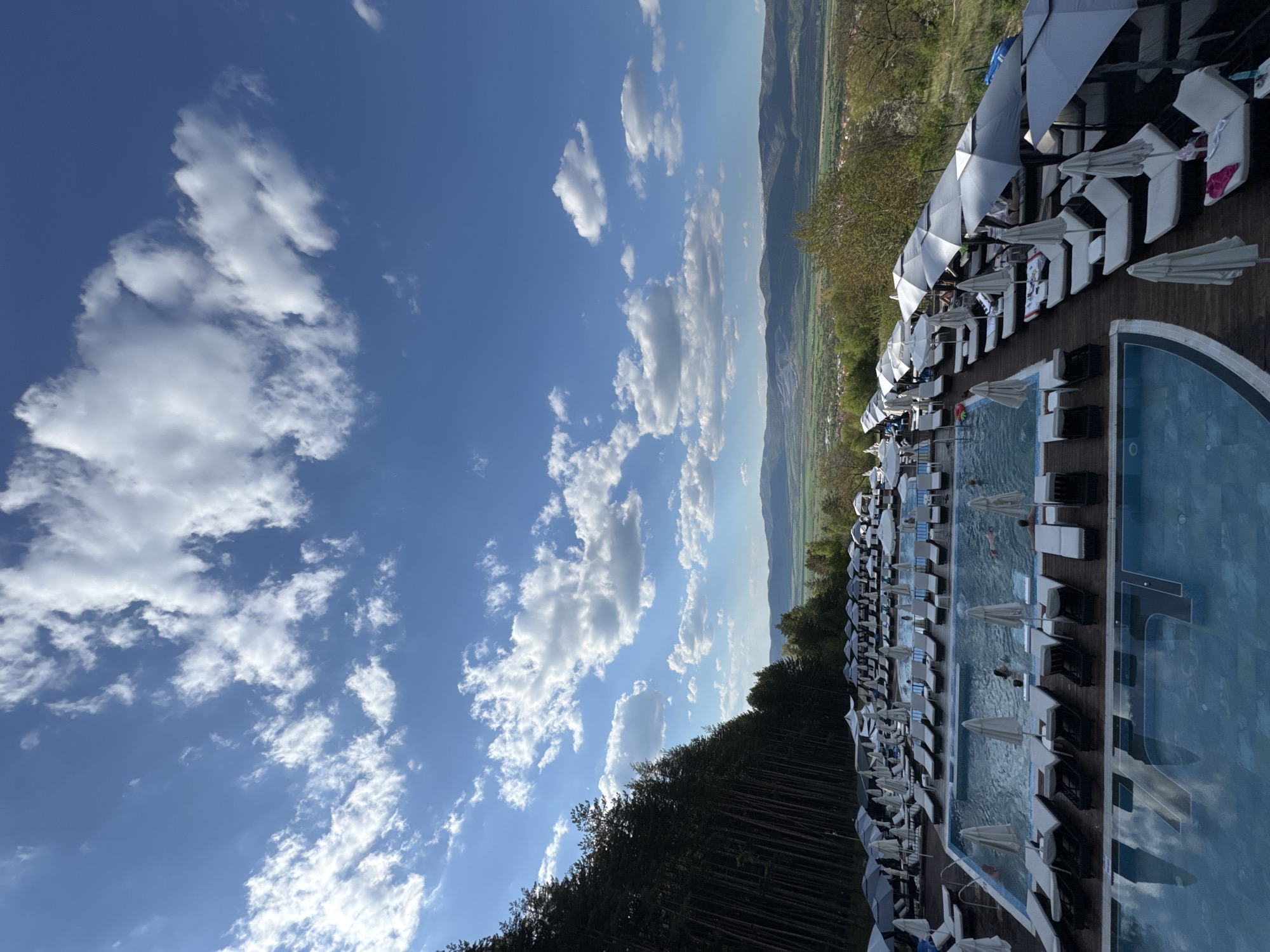 A long rectangular swimming pool surrounded by lounge chairs and white umbrellas is set against a backdrop of mountains and a partly cloudy blue sky. The location is likely a resort or hotel in Sapareva Banya, Bulgaria. The pool has people swimming in it. Trees border one side of the scene.