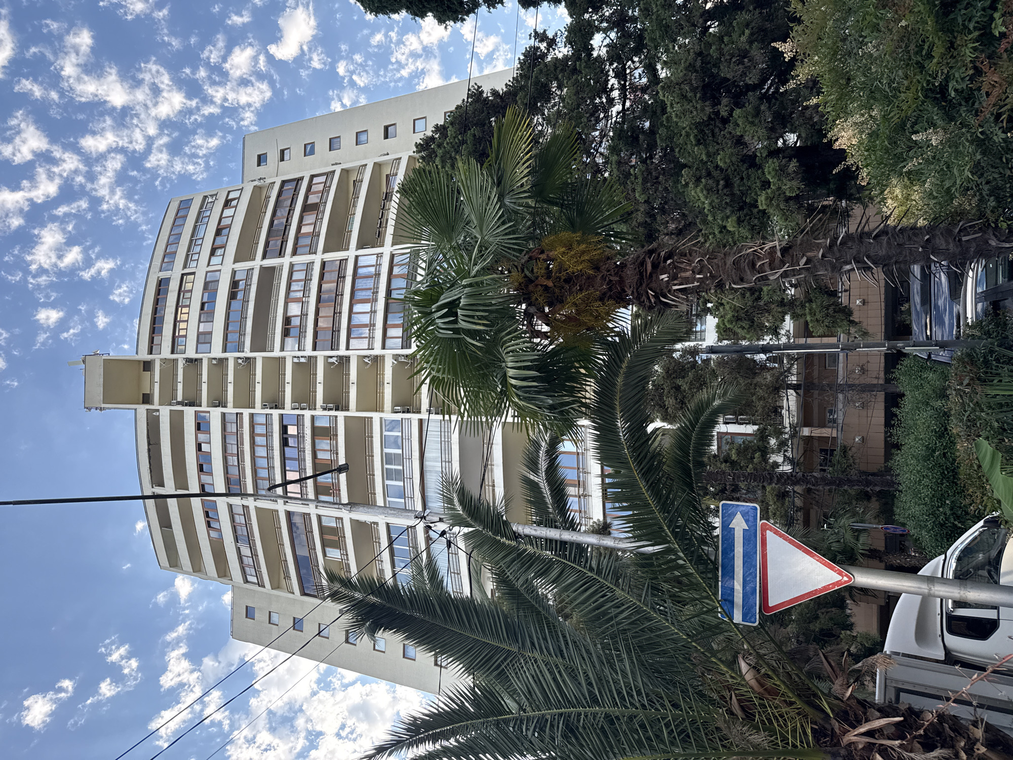 This media file depicts an outdoor scene with a modern apartment building and lush tropical vegetation under a partly cloudy sky. The building is tall and curvilinear, with many balconies and windows. Palm trees and other dense green foliage are in the foreground and surrounding the building, suggesting a warm climate. A road sign is visible, indicating traffic direction. The scene appears to be set in a developed urban area, possibly a resort town, with the presence of power lines and part of a vehicle. The lighting suggests it is daytime, and the weather appears pleasant with scattered white clouds against a blue sky. There is no visible text in the image. The overall impression is one of a sunny day in a vibrant, possibly coastal, environment.