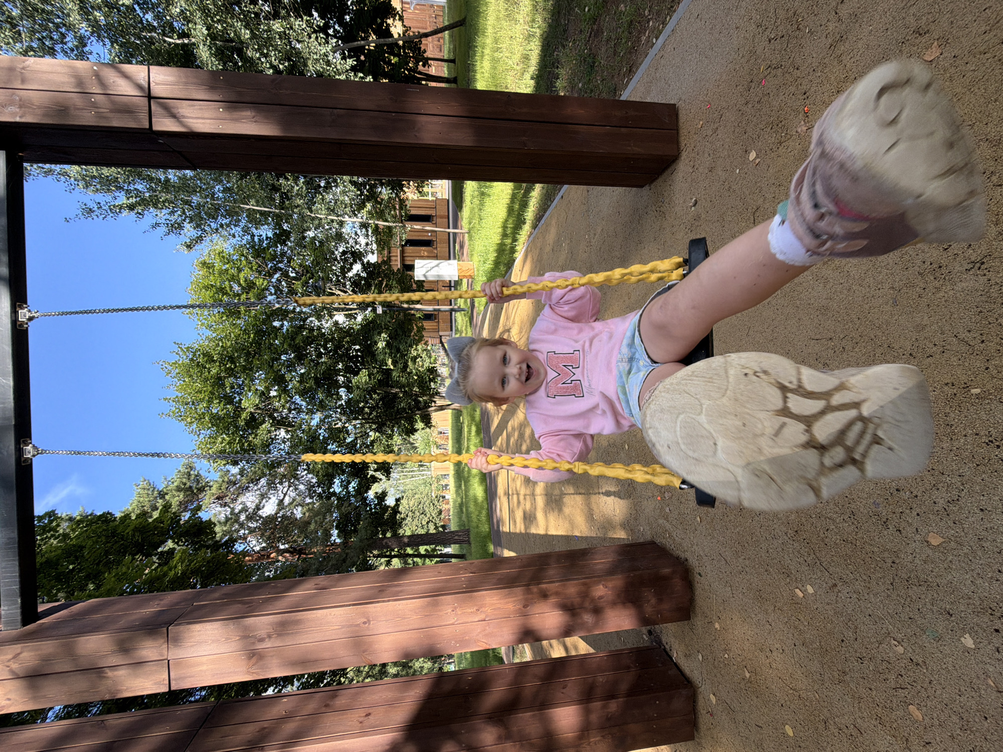 In Malakhovka, Russia, a young girl with grey cat ears on her head is joyfully swinging on a yellow rope swing. She wears a pink sweatshirt with a large 'M' on it and denim shorts. Her right foot is prominently in the foreground, mid-air with a worn sneaker. The swing set has two thick, dark brown wooden posts. The background features a bright blue sky, lush green trees, and a modern-looking wooden building. The ground is covered with sand, with scattered autumn leaves.