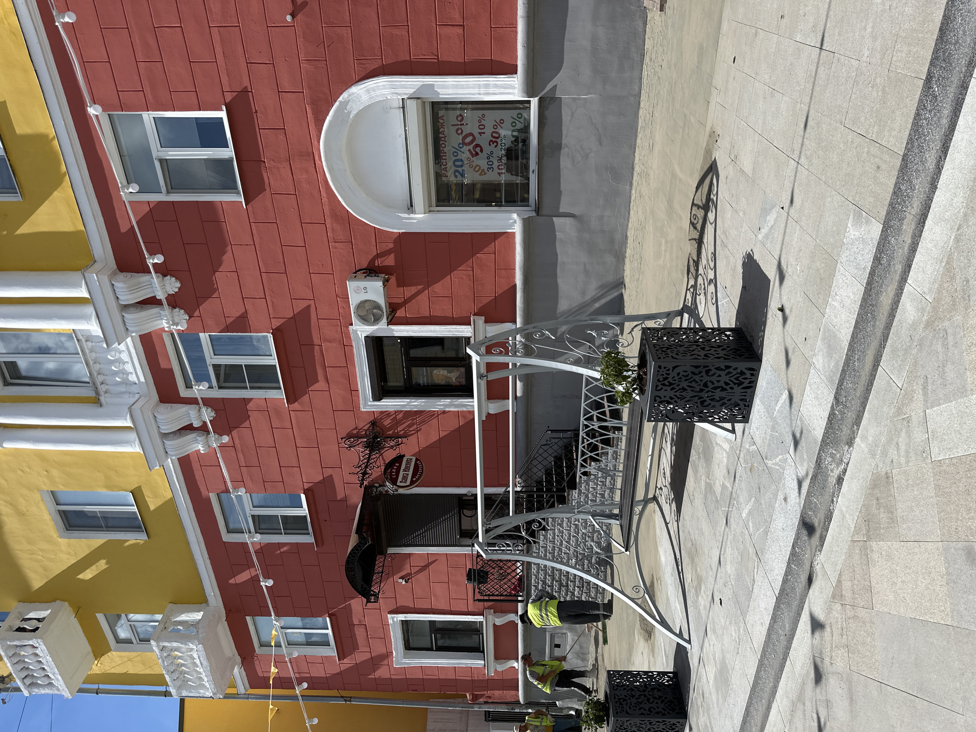 In Putyatino (Semivragovskaya S/A), Russia, a commercial building with a vibrant red facade is depicted under a clear sky. The building features yellow accents on its upper sections and white decorative elements around the windows and balconies. Two men in high-visibility vests are engaged in cleaning activities on the paved sidewalk in front of the building. One man is using a broom, and the other appears to be sweeping. A decorative metal swing is positioned on the sidewalk, and two large, ornate planters are placed on either side of it. A window on the red facade displays a "SALE" sign with various discount percentages. An air conditioning unit is mounted on the wall near one of the windows. The surrounding environment is a sunlit urban setting with paved walkways and shadows cast by the building and the swing.