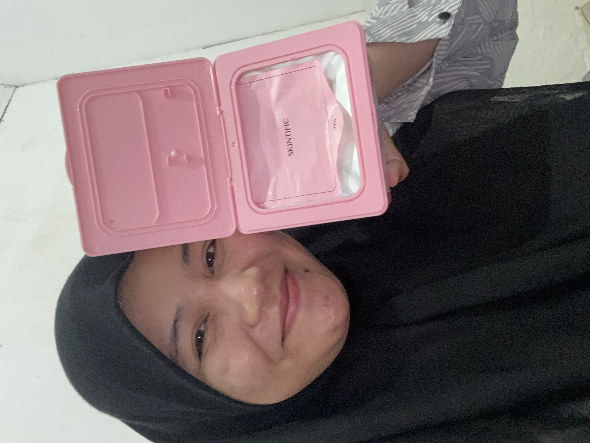 A woman wearing a black hijab and a patterned shirt is holding a pink container with a white cloth inside. The container is open, revealing the cloth. The woman is smiling and looking at the camera. The background is a white wall. The image appears to have been taken indoors. The woman's location is not specified, but the user provided the city of Banjarbaru, Indonesia.