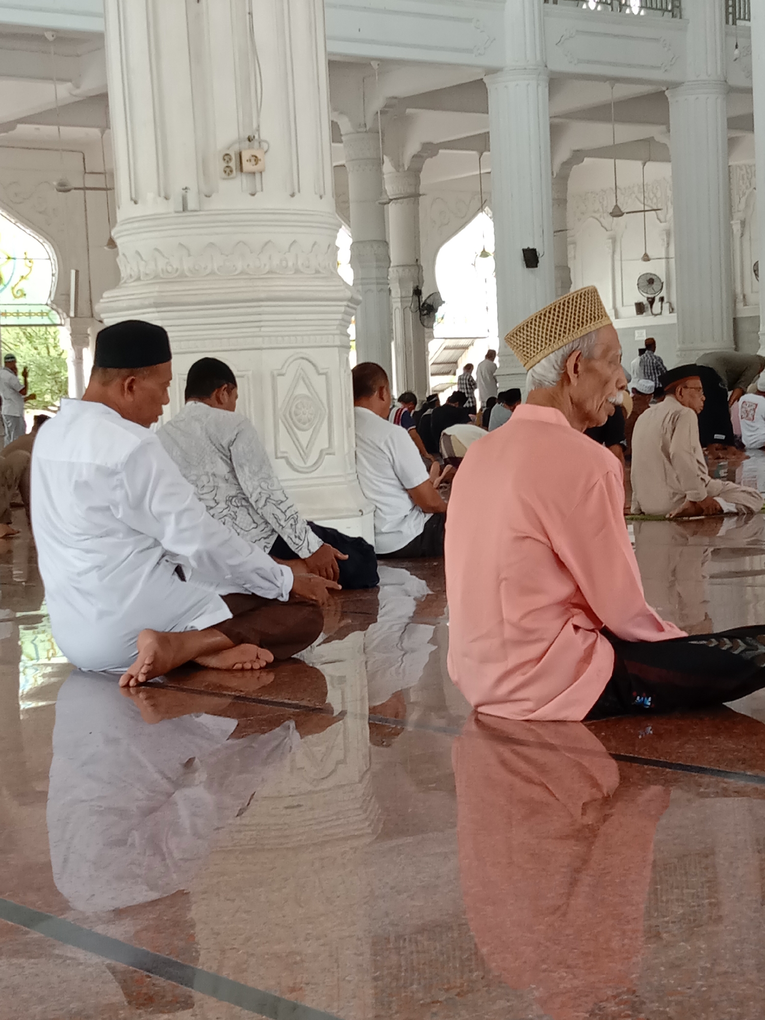 In the city of Lhokseumawe, Indonesia, inside a mosque, several men are seen sitting on the polished floor, some in prayer. The interior features large white columns with intricate carvings and a high, white ceiling with visible ceiling fans. Natural light streams in from the sides, illuminating the space and reflecting off the glossy floor. In the foreground, an elderly man with a white mustache wears a patterned gold cap and a light salmon-colored tunic, seated with his legs crossed. To his left, another man in a white tunic and black pants is also seated, facing away from the camera. Further back, other men are visible, some in white attire, others in darker clothing, some leaning forward as if in prayer or contemplation. Electrical outlets and wires are visible on one of the columns. The overall atmosphere appears to be one of quiet devotion and community gathering within the mosque.