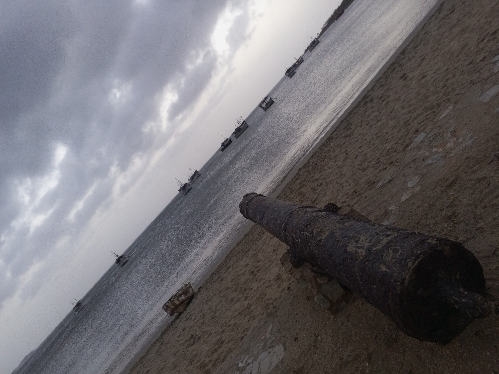 The image displays a diagonally composed coastal scene under a heavily overcast and cloudy sky. A strong diagonal line, defined by the shoreline and a series of boats, runs from the bottom right to the upper left of the frame.
In the immediate foreground, a large, dark, and heavily weathered cannon rests on sandy ground, its barrel pointing towards the water. The cannon appears to be made of aged iron, showing significant corrosion and texture from prolonged exposure to the elements, underscoring its historical nature. The ground around it consists of light brown sand mixed with scattered stones.
The midground is dominated by a calm body of water, likely a bay or sea, which exhibits subtle ripples on its surface, reflecting the muted grey light from the sky. Scattered across this water are numerous small to medium-sized boats, appearing to be fishing vessels or utility boats, each with various structures like small cabins and masts. They are sparsely distributed and extend into the distance along the coast, suggesting a maritime community.
The sky is completely covered with dense, grey clouds, indicating a very overcast day with diffused light. A faint, low-lying landmass or distant shoreline is barely visible on the horizon towards the far left, blending into the cloudy atmosphere. There are no people visible in the image, contributing to a quiet, contemplative atmosphere. The scene evokes a sense of history and the enduring presence of a maritime culture in a place like Juan Griego, Venezuela. No discernible text is present in the media file.