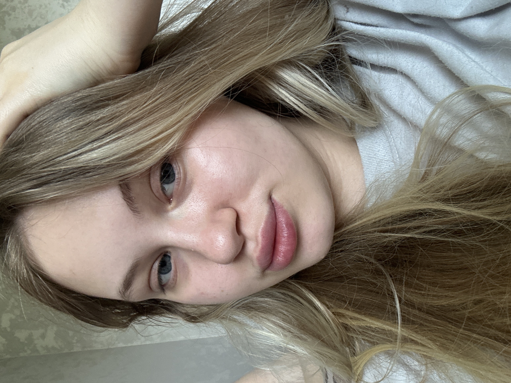 A young woman with blonde hair is taking a selfie in her home. She is wearing a grey t-shirt and her hand is resting on her head. The background appears to be a wall with a textured wallpaper pattern. The lighting suggests an indoor setting, likely daytime. There is no specific indication of the location within the city of Miass, Russia.