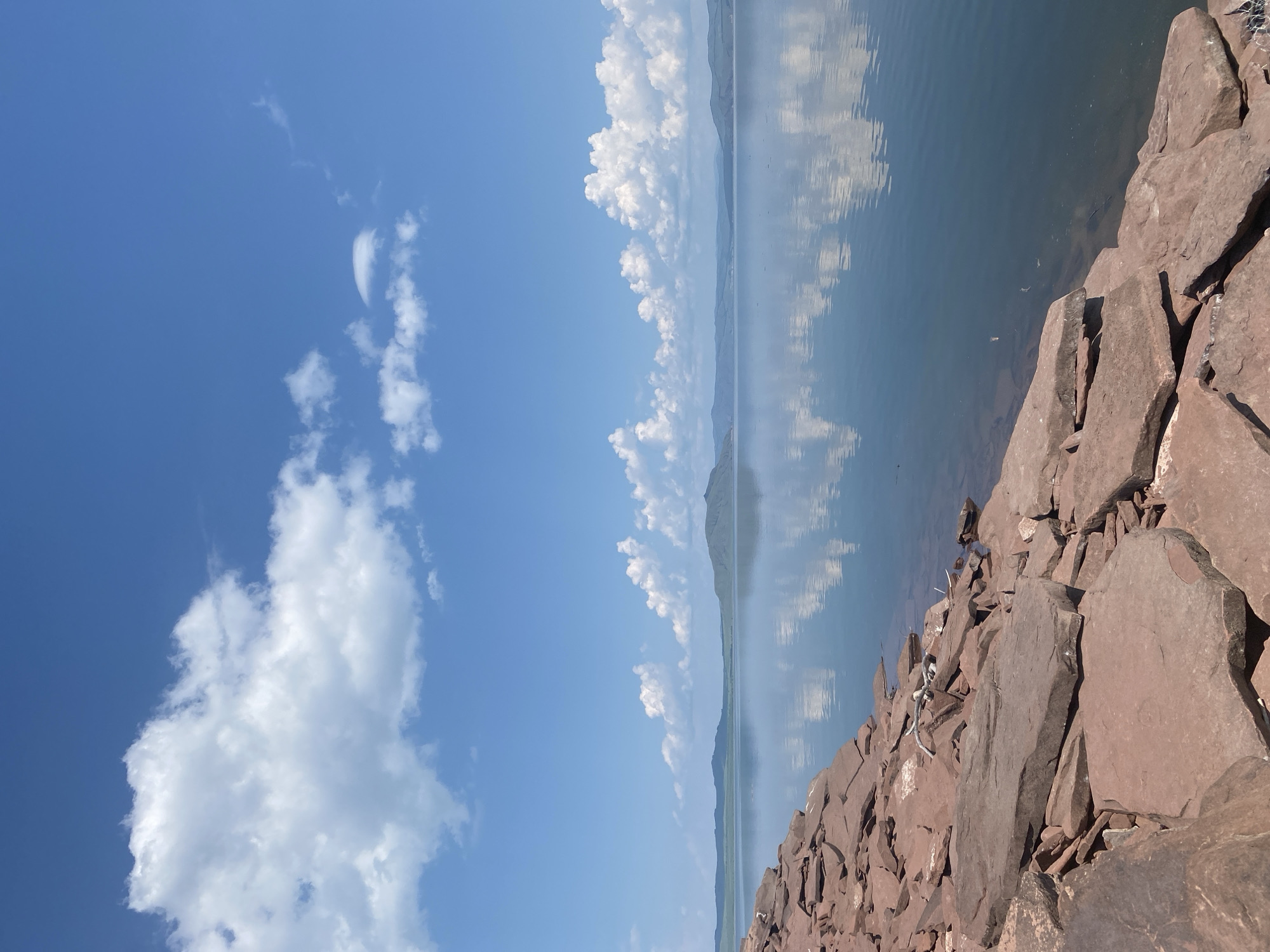The image depicts a serene natural landscape on a clear, sunny day in the vicinity of Ust'-Erba, Russia. The foreground is dominated by a rocky shore composed of large, reddish-brown stones, indicative of a natural, undeveloped coastline. The stones are unevenly scattered, suggesting a rugged terrain. Beyond the shore lies a vast, calm body of water, reflecting the bright blue sky and scattered white cumulus clouds. The water surface is smooth, creating a near-perfect mirror image of the sky and the distant, low-lying hills on the horizon. These hills are a muted green, softened by the distance and atmospheric haze. The overall impression is one of tranquility and natural beauty, with a sense of open space and a clear, expansive sky. There are no people or man-made structures visible in the scene.