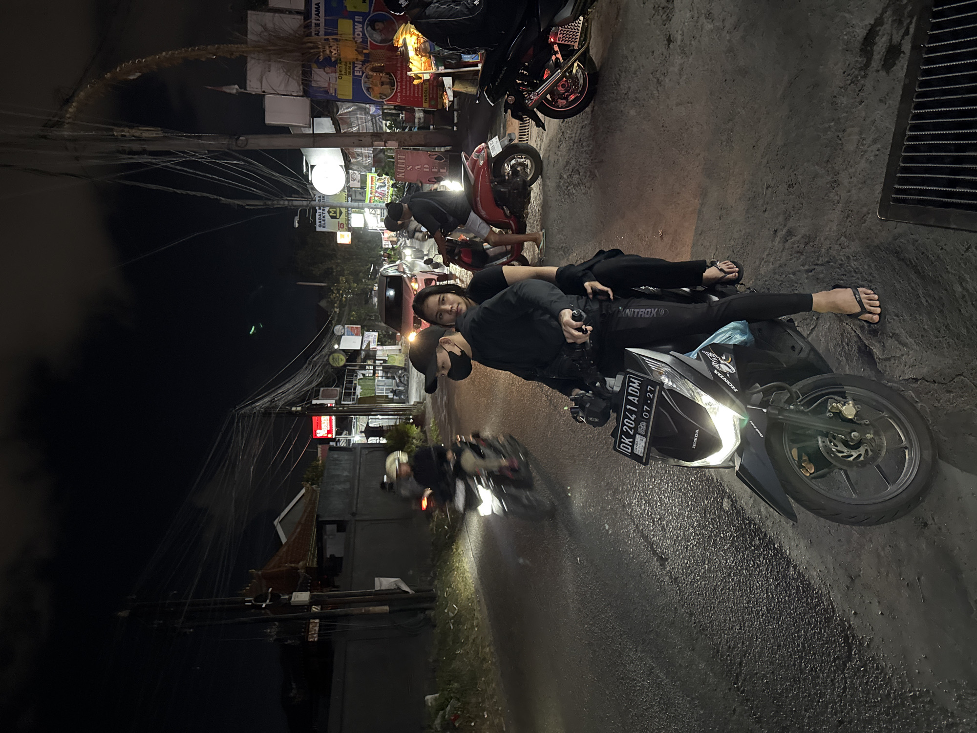This media file captures a nighttime scene in Denpasar, Indonesia, featuring two people resting on motorcycles. The subjects, a man and a woman, are casually positioned on their bikes, with the woman lying down and the man sitting. Both are wearing casual attire, including flip-flops. The environment appears to be a street or roadside, indicated by the presence of motorcycles, asphalt road, and overhead electrical wires. The time of day is clearly night, with artificial lighting from streetlights and signs illuminating the scene.
Several motorcycles are visible in the background, suggesting a busy street or a parking area. One motorcycle in the foreground has a license plate that reads "DK 2041 ADM" and a date stamp "07.27." There are also various signs in the background, some displaying text in a language that appears to be Indonesian. Visible text includes "HONDA," "KNITROX," "GADAI MGA ELEKTRIK," "ARIE PAMA," and "EN NOW !!". The road surface appears wet, hinting at recent rain or damp conditions. The overall mood seems relaxed, with the subjects taking a break amidst the urban night.