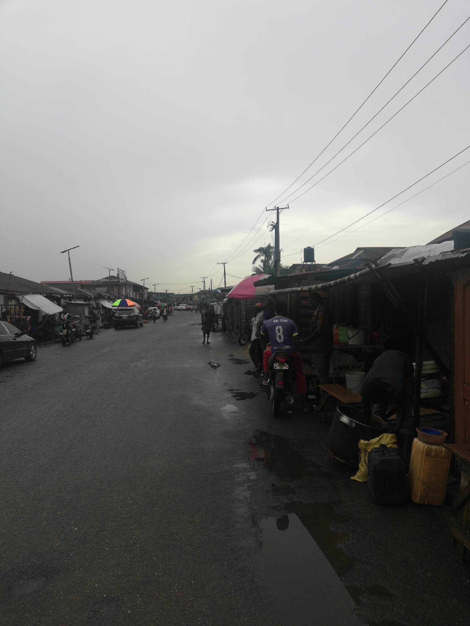 The image depicts a street scene in Lagos, Nigeria, on an overcast day. The street is wet, suggesting recent rain. The road stretches into the distance, lined with informal shops and structures on both sides. On the right side of the road, several men are congregated around a motorcycle and the shop, some appearing to be engaged in conversation or business. Various containers and buckets are visible near the shops. On the left, parked cars, motorcycles, and more informal shops are present. People are seen walking and moving along the street. Overhead power lines run along the length of the street. In the distance, more buildings and street activity can be observed.