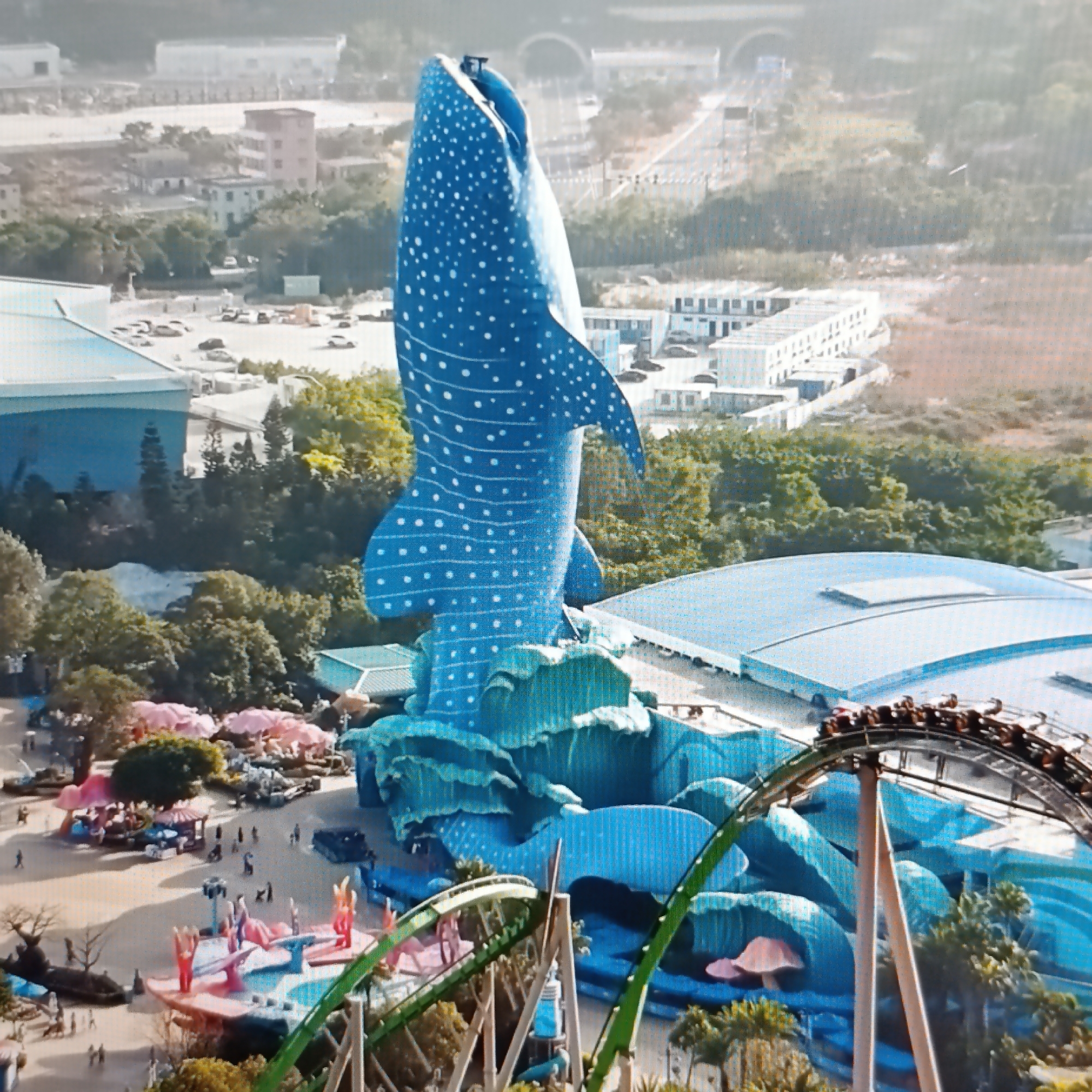 This aerial view captures a vibrant amusement park with a prominent blue whale shark sculpture as the central attraction. The sculpture is detailed with white polka dots and is surrounded by turquoise, wave-like structures. In the foreground, a roller coaster track curves through the scene, with a train of riders visible at its apex. The park area below the sculpture is populated with people, colorful mushroom-shaped stalls, and various rides. Further in the background, buildings and roads indicate a semi-urban environment. The image does not depict the city of Dubăsari town council, Moldova; it appears to be an international location.