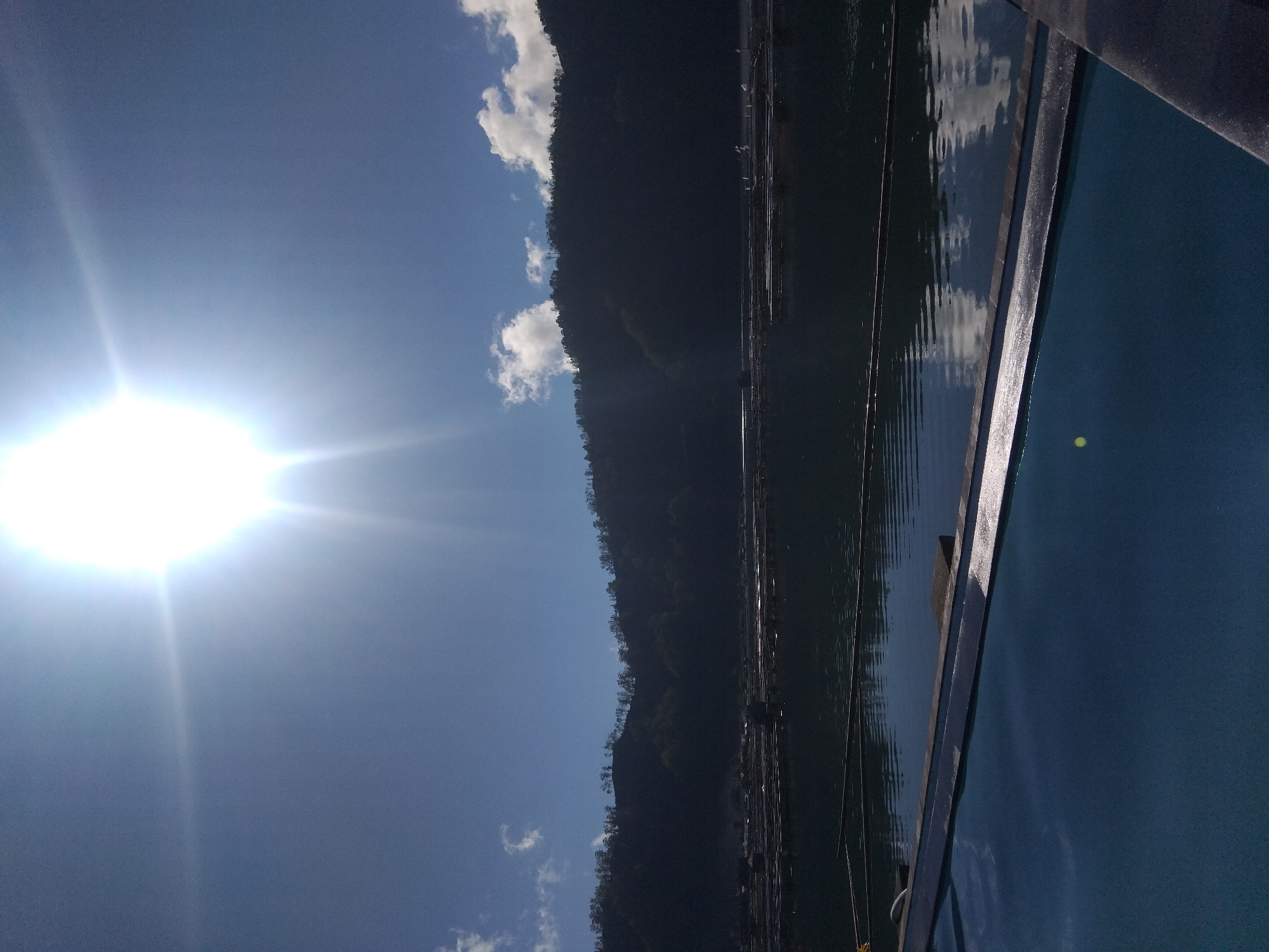 The image depicts a body of water reflecting a mountain range under a bright sun. A portion of a boat is visible in the foreground. The location is specified as Unknown, Unknown. The sky is blue with a few scattered clouds. The water surface reflects the sky and the mountain, creating a mirrored effect.