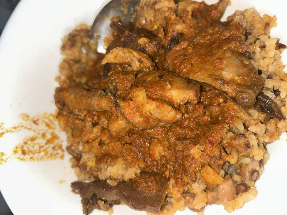 A white bowl contains a serving of food. The base layer consists of a granular mixture, appearing to be rice and beans, with a light brown and beige coloration, interspersed with darker bean specks. This base is covered by a thick, reddish-orange stew, rich in texture and color, indicative of a tomato and chili pepper base. Embedded within the stew are various pieces of protein, including irregular chunks of lighter-colored meat, possibly poultry or red meat, and darker, elongated pieces that may be dried fish or offal. A silver-colored spoon is partially submerged in the food on the right side of the bowl. Splatters of the reddish-orange stew are visible on the upper interior rim of the white bowl. No people or other environmental details are visible.