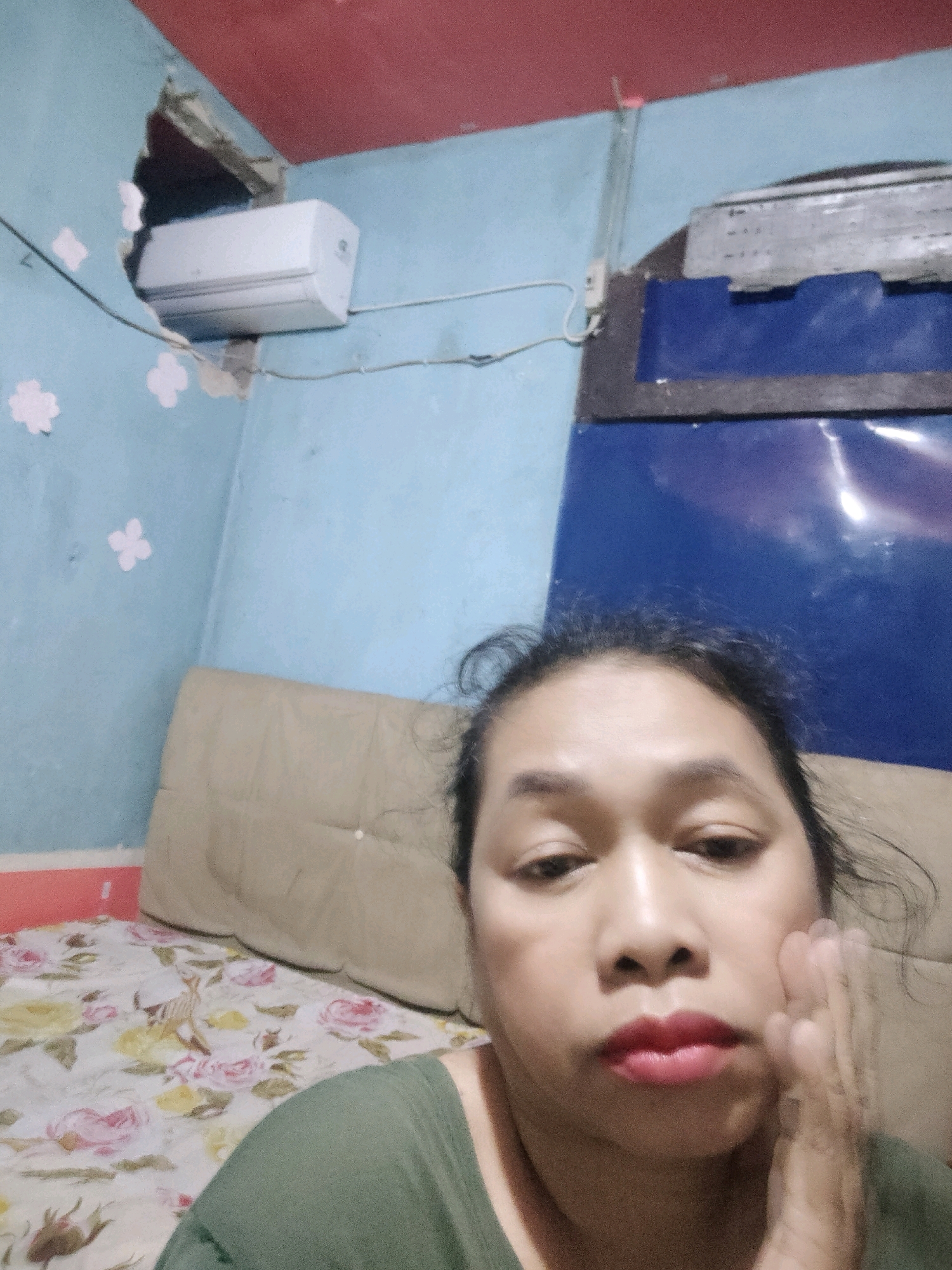 A woman is taking a selfie in a room. The room has a light blue wall with a hole in the corner near the ceiling, where an air conditioner is installed. The ceiling is red, and there is a blue cabinet with a silver-colored object on top in the background. The woman is wearing a green shirt and has her hand on her cheek. In the foreground, a bed is visible with a floral-patterned sheet and a beige headboard. The location is not specified in the image.