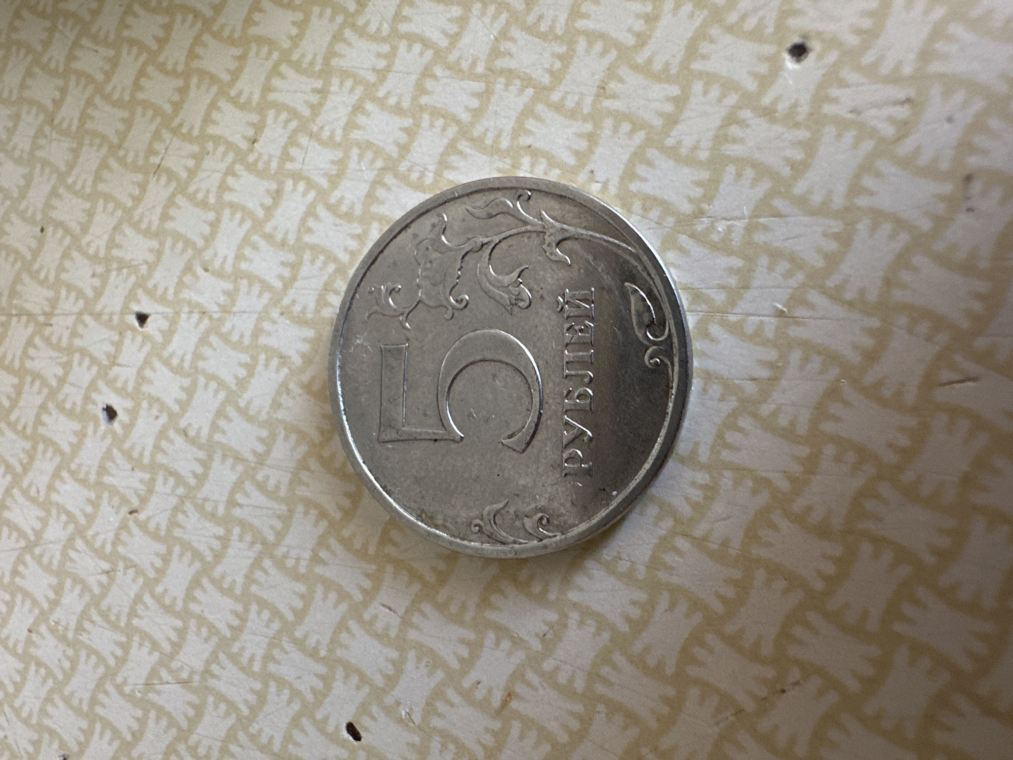 A 5 ruble coin is resting on a light-colored surface with a repeating pattern of stylized white figures resembling praying hands. The coin is silver in color and is oriented with the denomination "5" facing upwards and slightly to the right. The Cyrillic text "РУБЛЕЙ" (RUBLEY) is visible below the number 5, indicating the currency. Decorative floral elements are also present on the coin's surface. There are a few small dark specks and faint lines on the background surface, possibly dust or imperfections. The image is a close-up shot, and no specific location in Biysk, Russia, can be determined from the provided visual information.