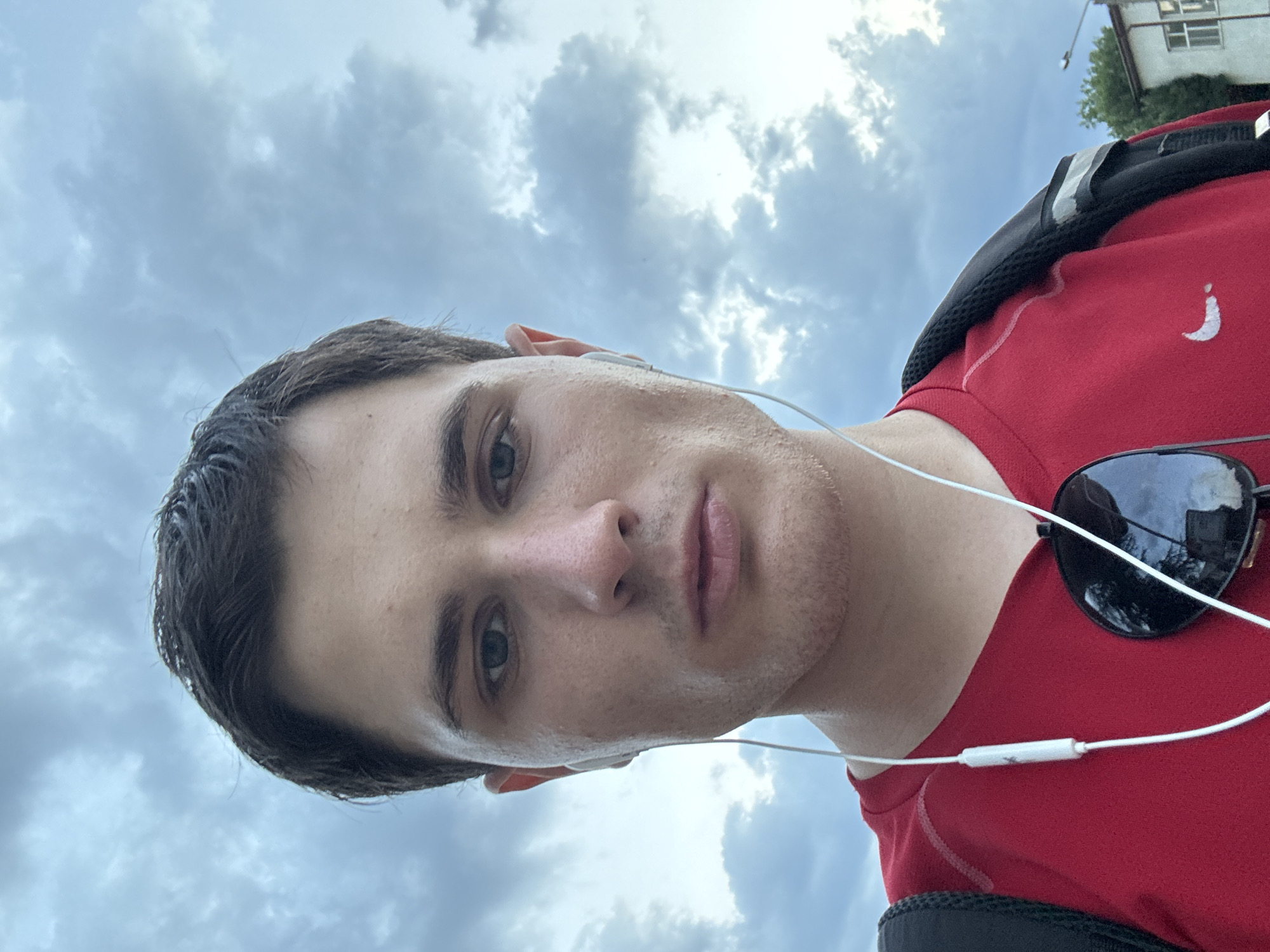 A close-up, outdoor selfie shows a fair-skinned, young adult male with short, dark hair and light blue eyes. He is wearing a red t-shirt with a white Nike logo, white earbuds, and sunglasses hanging from the collar of his shirt. He is looking directly at the camera with a neutral expression. The background is a cloudy sky with hints of greenery and a building visible on the right side of the frame, suggesting an urban environment. The lighting indicates it is daytime. The location is identified as Stara Zagora, Bulgaria.