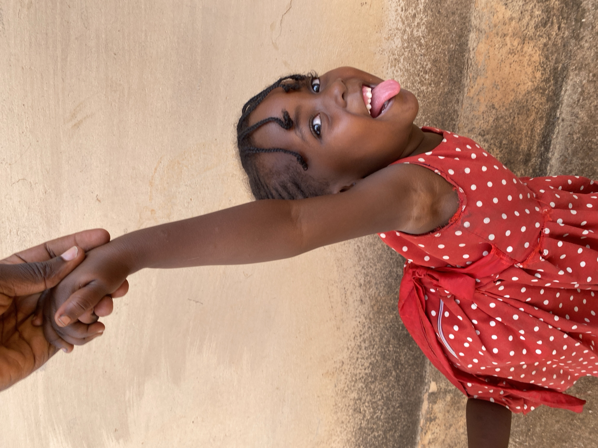 The image captures a joyful and playful moment featuring a young Black child and an adult, set against a light brown, textured wall in Kuru Dungus, Nigeria. The child, positioned diagonally across the frame and seen from the chest up, looks directly at the viewer with a wide smile and her tongue playfully sticking out. Her hair is styled in neat braids. She is wearing a vibrant red dress adorned with white polka dots. An adult's hand, partially visible on the left side, firmly holds the child's outstretched left hand, suggesting a close connection. The background consists of a light-colored, somewhat rough wall surface, with a darker, more textured ground visible in the lower right, indicating an outdoor or semi-outdoor setting under natural daylight. The overall mood of the image is lighthearted and cheerful. There is no visible text.