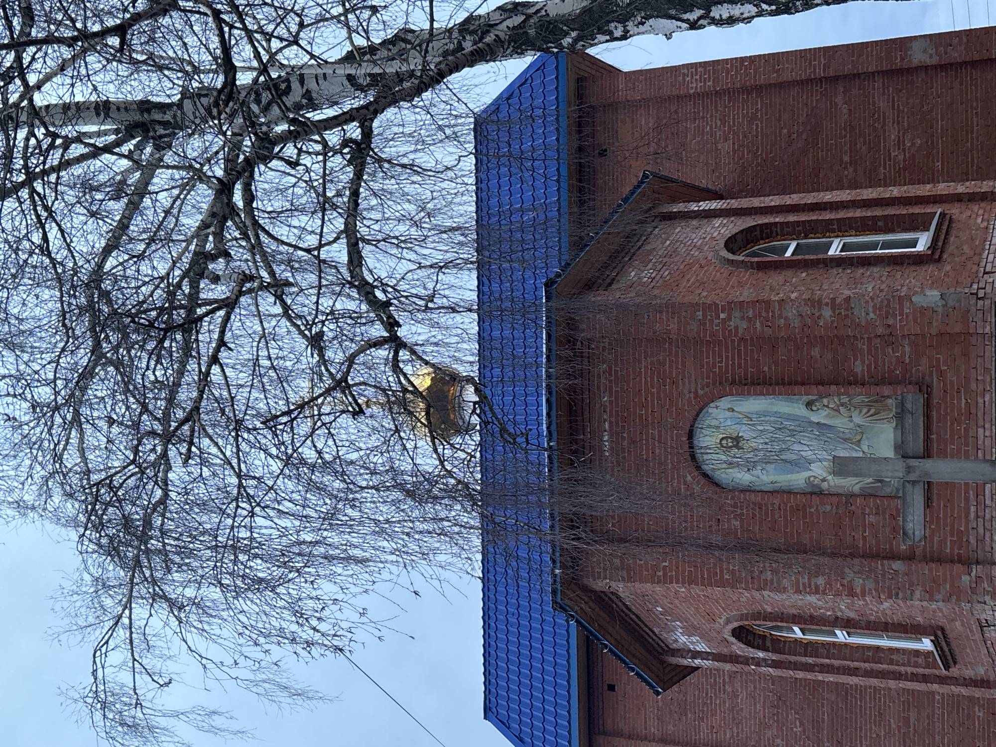 A brick church with a blue tile roof stands in Фоки, Russia. Bare tree branches fill the left of the frame, partially obscuring a gold dome. The church's brick facade has arched windows and a religious mural above a wooden cross. The sky is pale blue.