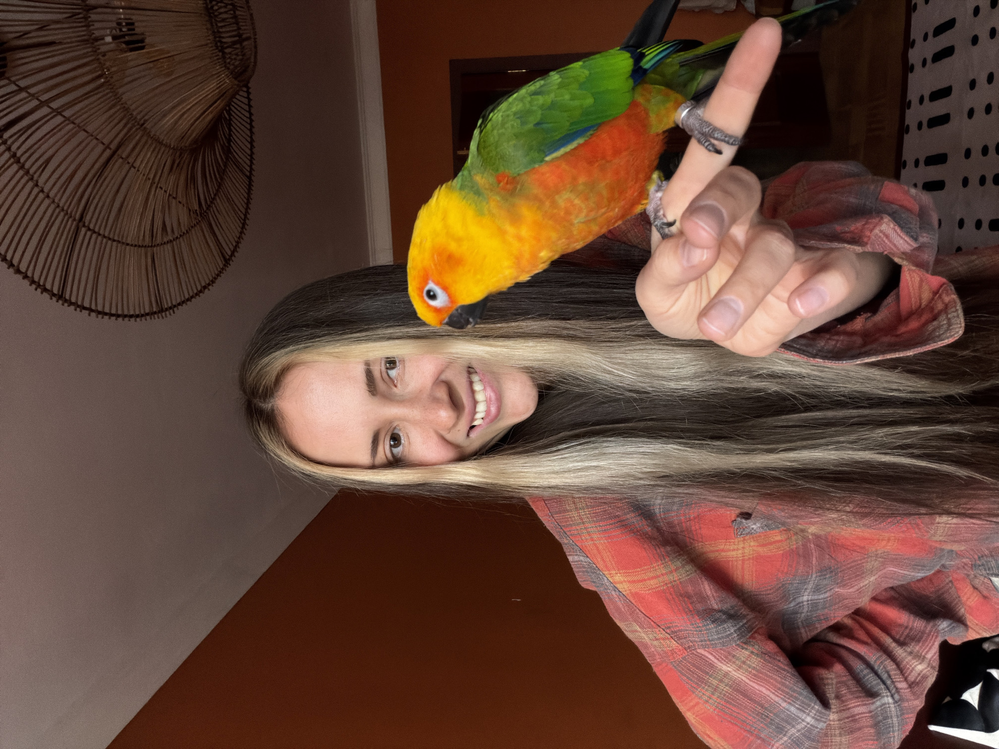 A woman smiles at the camera, turned sideways. She has long dark and blonde hair and wears a red and tan plaid shirt. A colorful parrot, primarily yellow, orange, and green, perches on her finger. A decorative wicker lamp shade is visible in the background, along with sections of a light brown and dark brown wall and a black and white patterned surface. There is no evidence of Khimki, Russia in the image.