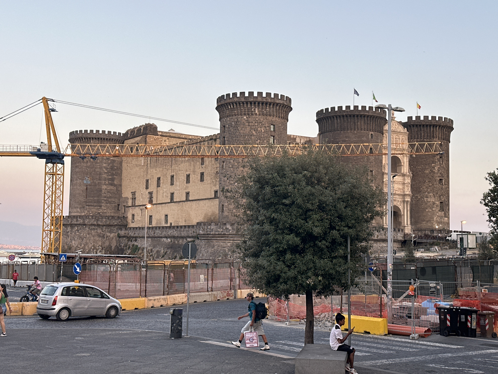 The image shows a wide shot of Castel Nuovo, a medieval castle in Naples, Italy. The castle, with its distinctive five towers and crenellated battlements, is the central focus. In front of the castle, a construction site is visible, with orange safety netting, cranes, and barriers. Several people are present: a woman in a green top walks in the foreground to the left, a silver car is parked on the street, and a man with a backpack walks across the street, holding a shopping bag. Another young person sits on a ledge in the lower right corner, looking at their phone. The sky is a pale, clear blue, suggesting it's either early morning or late afternoon. The overall scene captures a blend of historic architecture and ongoing urban development in Naples.