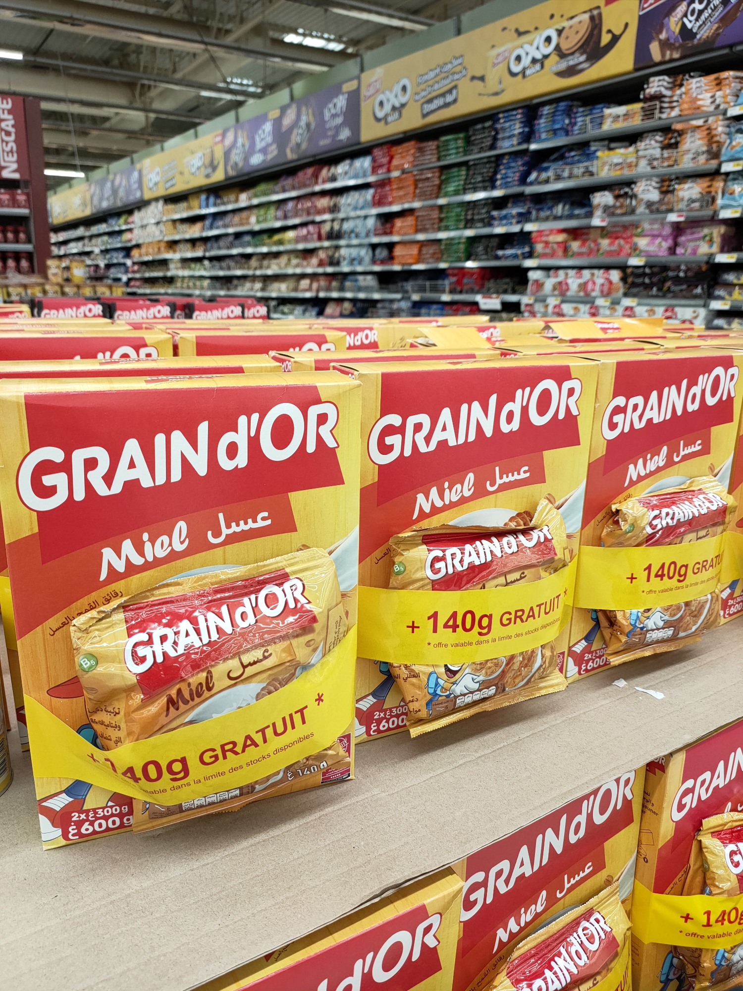 The image depicts a retail environment, specifically a supermarket aisle in Cebalat, Tunisia, displaying boxes of "GRAIN d'OR" cereal. The cereal boxes are predominantly yellow and red, with prominent "GRAIN d'OR" branding. Many of the boxes feature promotional bands indicating "+ 140g GRATUIT" (free). Some individual cereal packages are visible within the boxes, also branded "GRAIN d'OR Miel" (honey). The background shows shelves stocked with various products, suggesting a well-stocked store. The lighting appears to be artificial, typical of an indoor retail space. No people are visible in the image. The overall scene is one of commercial display and product merchandising.