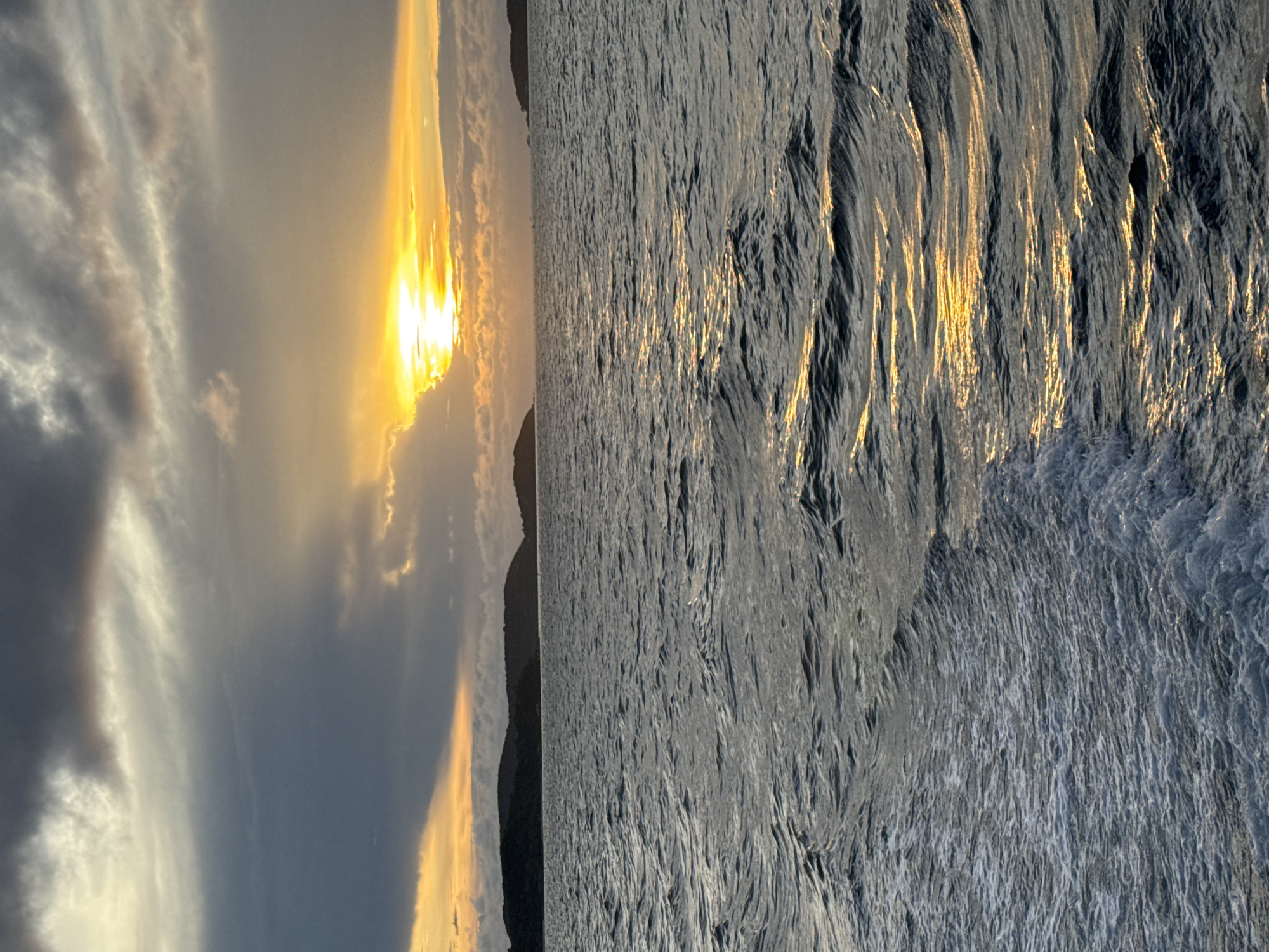 The image depicts a sunset over the ocean in Pattaya City, Thailand. The sun is setting behind a silhouetted landmass, casting golden light through the clouds and reflecting on the surface of the water, which displays textural wave patterns.