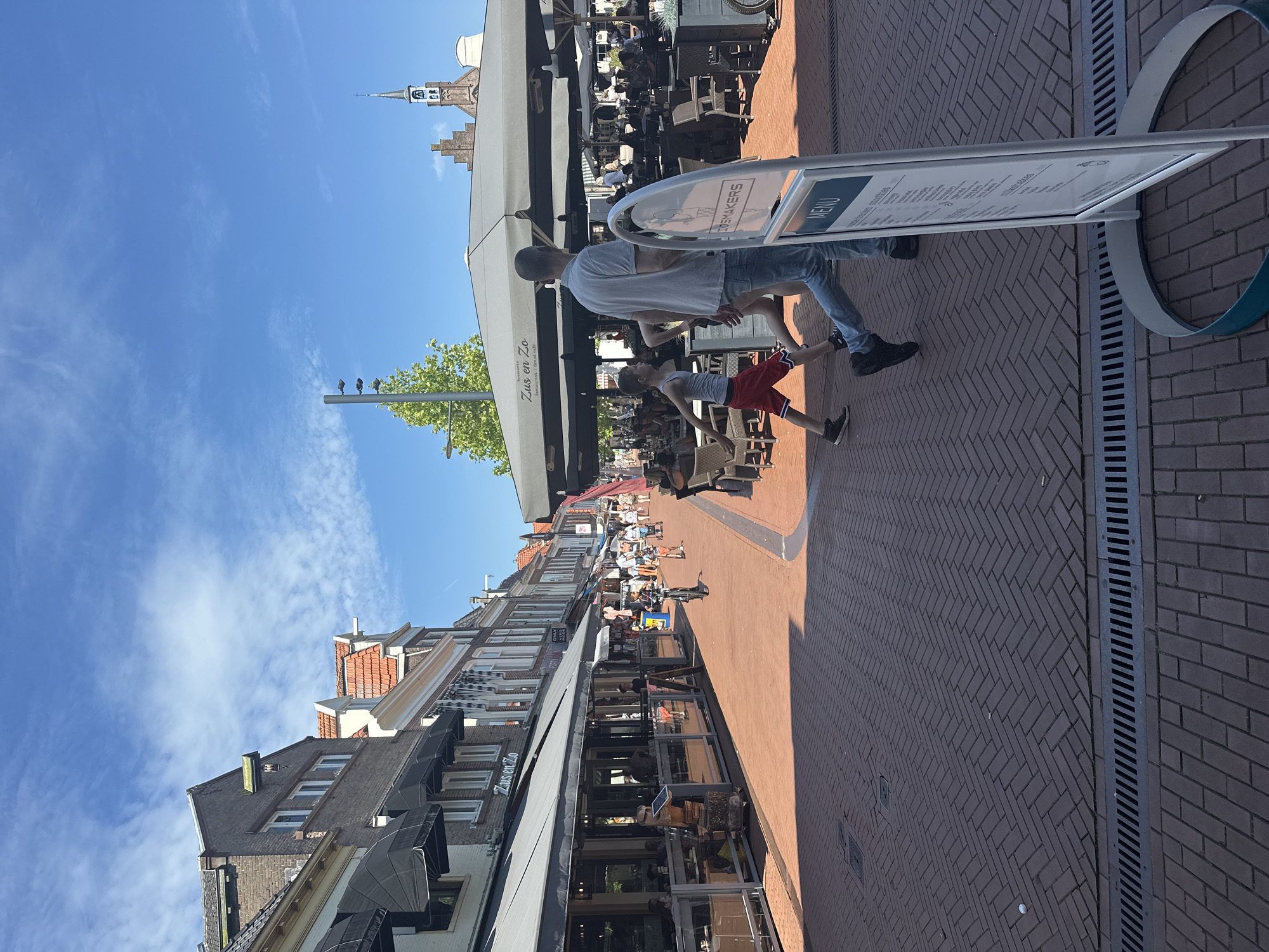 This image depicts a street scene in Steenwijk, Netherlands, likely a town center with shops and restaurants. The foreground is dominated by a red brick paved street laid in a herringbone pattern, with a metal drain grate running horizontally across the lower portion of the frame. A white A-frame sign with a menu is prominently positioned on the right, displaying the word "MENU" and illegible text.
In the middle ground, a man and a young boy are walking from right to left. The man is wearing a light blue t-shirt and jeans, and the boy is wearing a grey tank top and red shorts. They are walking past an outdoor seating area of a restaurant, which features large umbrellas and tables with chairs. Further down the street, other pedestrians are visible, some walking and others appearing to be dining or shopping. Buildings line both sides of the street, with awnings extending over storefronts. One awning has the text "Zus en Zo" visible. A church steeple with a clock can be seen in the distance on the right. The sky is blue with scattered white clouds. The overall impression is a bustling urban environment on a sunny day.