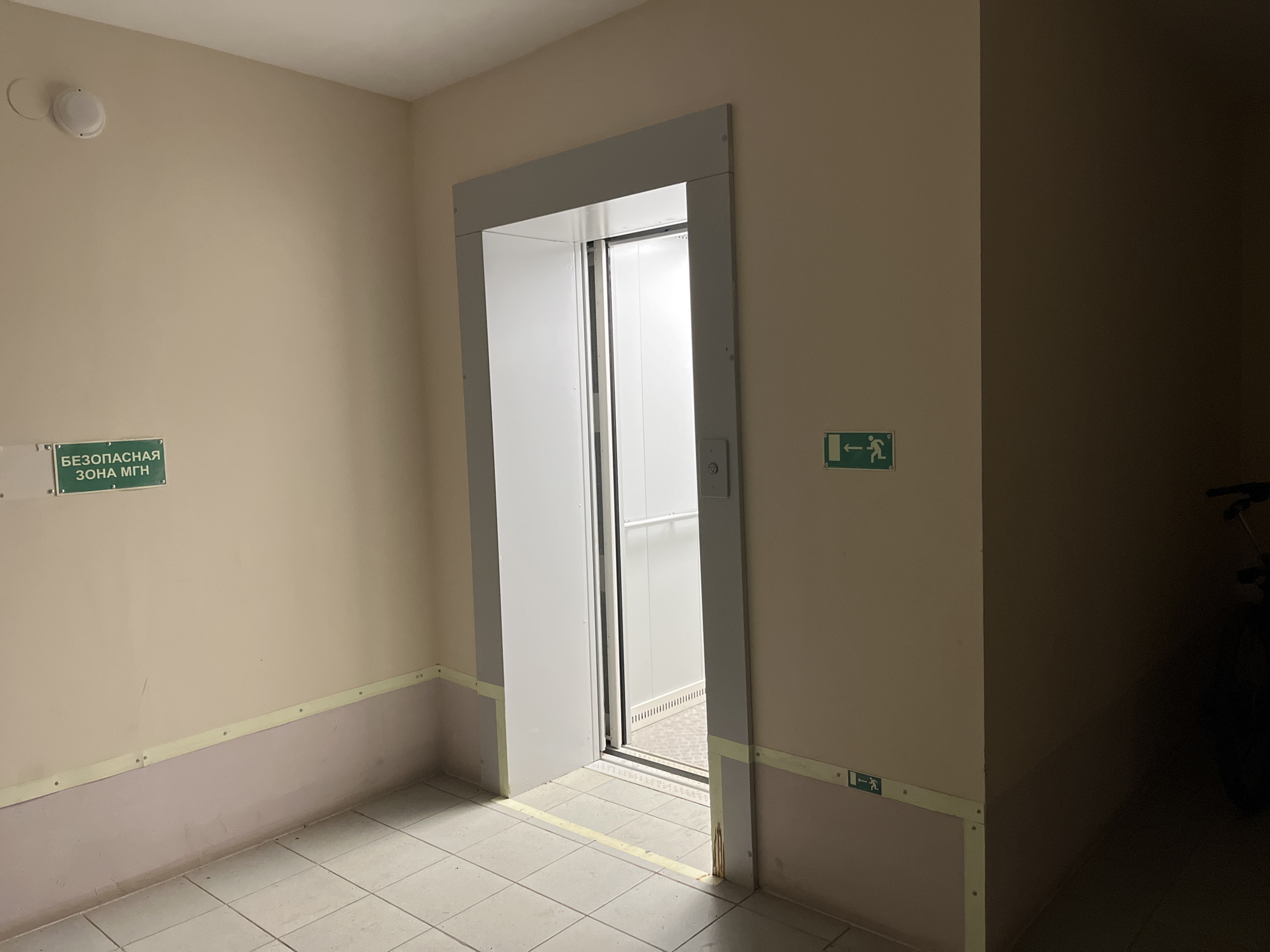 A photograph taken in Chaikovsky, Russia shows a hallway leading to an elevator. The elevator door is ajar, revealing the interior of the elevator car. The hallway walls are beige with a purple wainscoting detail. A sign in Cyrillic script reads "Безопасная зона МГН" which translates to "Safe zone for people with limited mobility." An emergency exit sign is also visible. The hallway floor is tiled in a grid pattern. A dark space is visible to the right of the elevator with a bicycle parked inside.