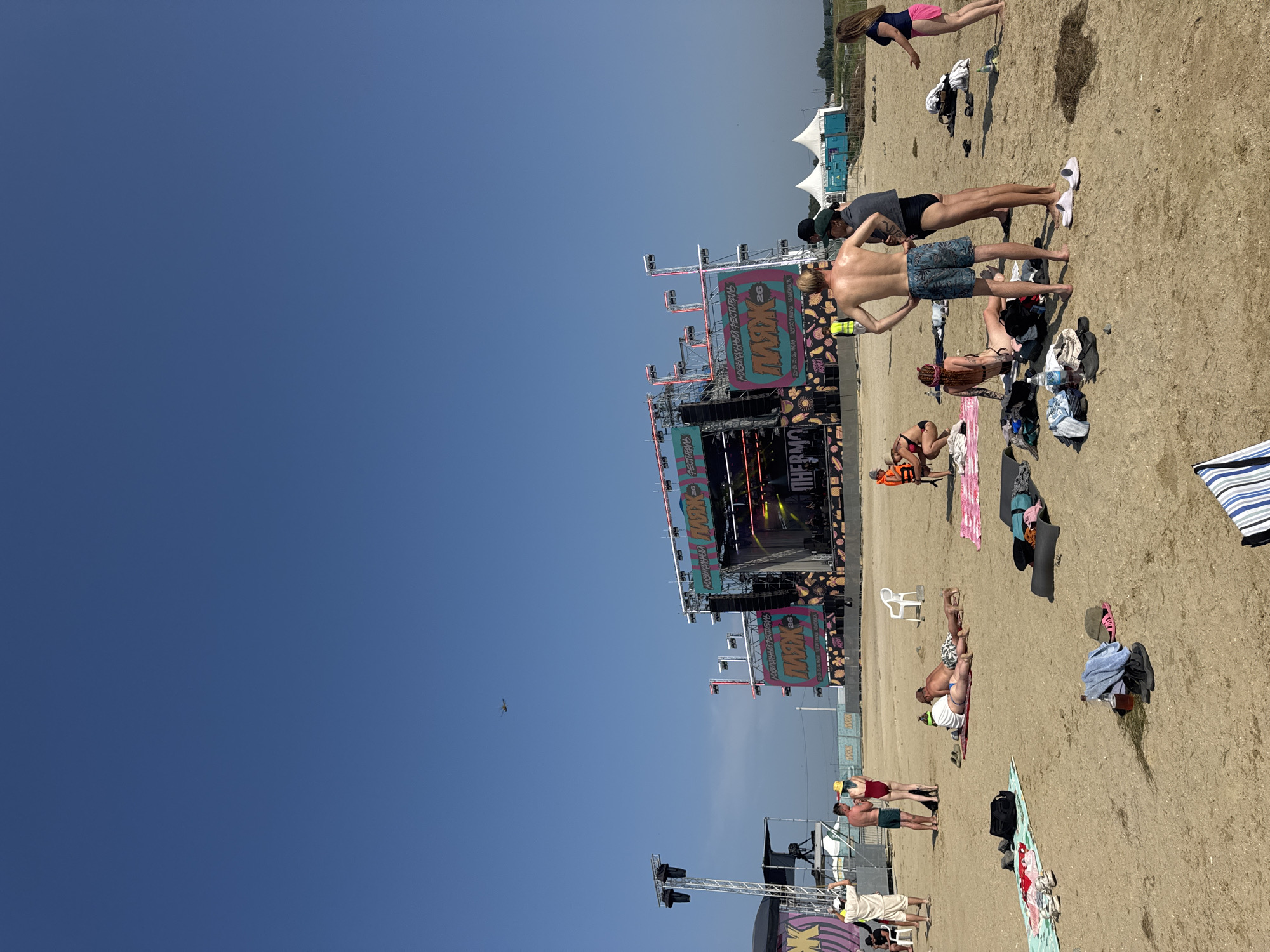 A wide shot shows a beach festival scene under a clear blue sky. A large stage structure with colorful banners reading "ПЛЯЖ 26" and "МУЗЫКАЛЬНЫЙ ФЕСТИВАЛЬ" dominates the middle ground. To the left, another stage structure with lighting rigs is visible. A few people are scattered on the sandy beach, some are sunbathing on towels, while others are standing and talking. In the distance, a helicopter is flying. In the background, white tents and some greenery suggest a remote location. The overall atmosphere is relaxed and summery, with an ongoing music festival.
