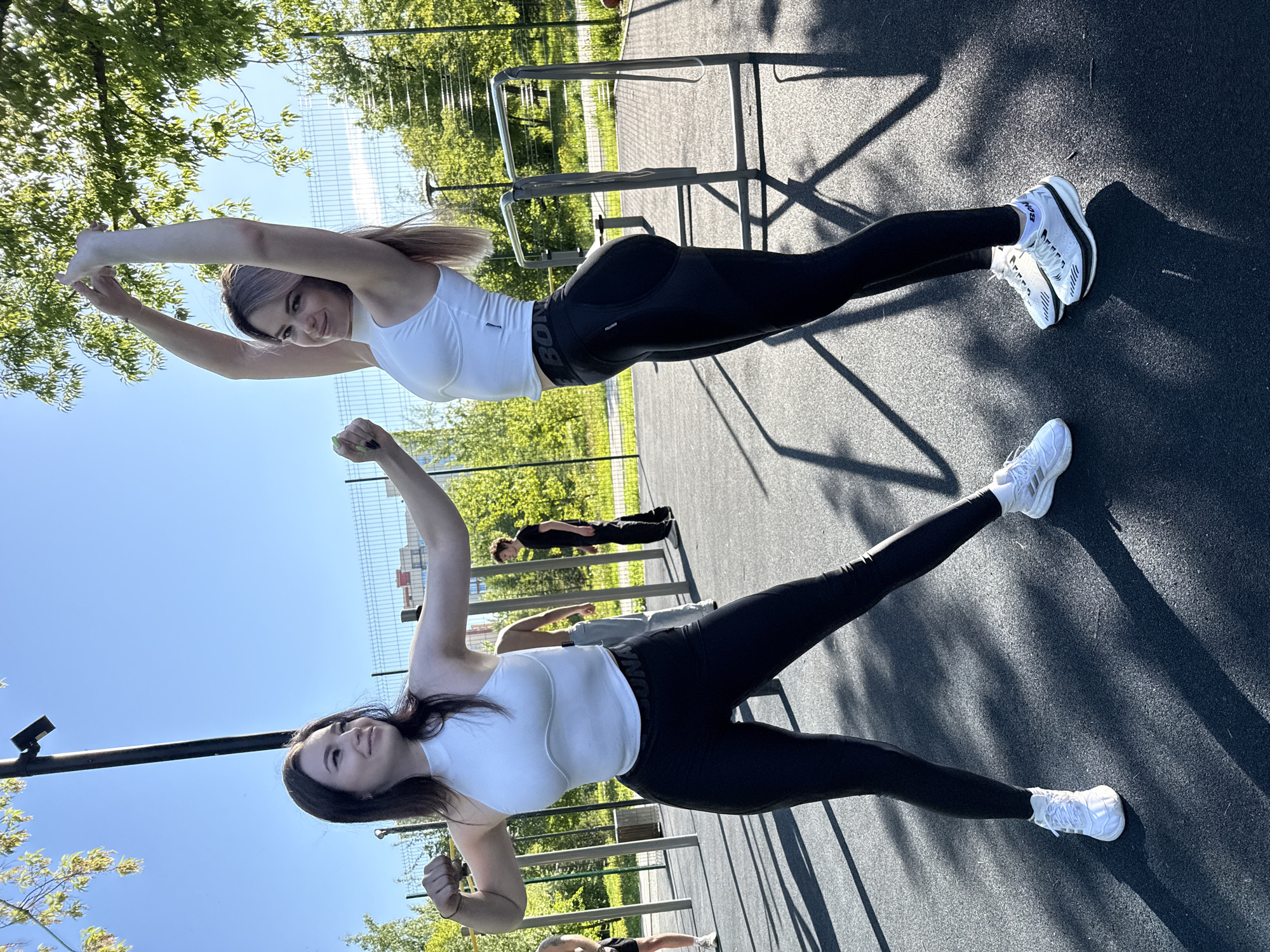 The image shows two women outdoors, presumably at a public workout area in the city of Unknown, Unknown. The woman in the top half of the image has blonde hair, is wearing a white tank top and black leggings, and has her arms raised above her head. Her legs are fully extended on the ground. The other woman is positioned in the bottom half of the image. She has dark hair and is also wearing a white tank top and black leggings. Her arms are above her head, in a similar pose to the other woman. Both are wearing white sneakers. The background contains workout equipment, trees, and other people further away. The ground appears to be made of a gray material, with shadows cast across it. The sky is clear and blue.