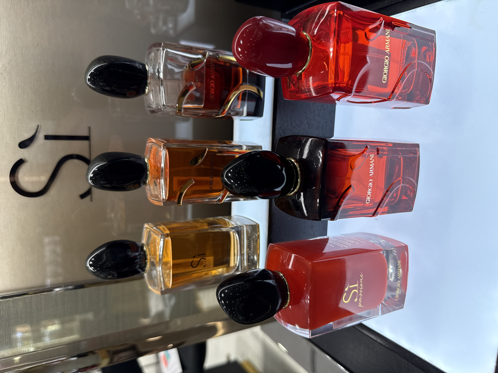The image displays a collection of Giorgio Armani Si perfumes arranged on a white display shelf. There are six bottles in total, with three in the back row and three in the front row. The bottles in the back row have amber-colored liquid and black caps, with the Giorgio Armani logo visible on the front. The bottles in the front row are red, with one featuring the "Si Passione" label and a gold Giorgio Armani logo, and the other two also bearing the Giorgio Armani logo and a red cap. The display is illuminated from below, creating a bright and reflective surface. The background appears to be a store environment, possibly a department store or a perfume shop, with a metallic backdrop featuring the "Si" logo prominently. There are no people visible in the image, and the scene is static, suggesting a product display. The location is identified as Astana, Kazakhstan.
