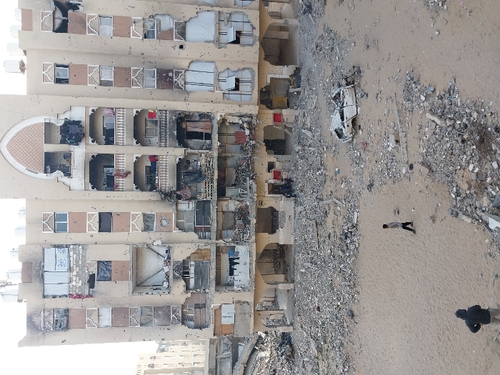 The image depicts a scene of extensive damage and destruction, centered around a multi-story building and an adjacent expanse of sandy ground covered in rubble.
The primary subject is a large, multi-story building, likely an apartment complex or residential structure, with a light-colored, possibly beige or cream, facade. It shows severe structural compromise across multiple floors. Many windows are shattered, blown out, or completely missing, revealing the interiors of numerous apartments. Large sections of the external walls and balconies are either collapsed, partially destroyed, or heavily scarred. Inside the exposed units, debris, damaged furniture, and what appear to be personal belongings, such as clothes hanging from former balconies, are visible. Some areas within the building appear blackened, suggesting fire damage. The building's exterior also exhibits numerous smaller impact marks, resembling bullet holes or shrapnel damage, spread across its facade. An ornate, arched architectural detail is visible on an upper-middle section of the building.
Adjacent to the building, to its right, is a wide area of sandy ground. This ground is extensively covered with large amounts of rubble, including chunks of concrete, twisted metal, and other debris from destroyed structures. Amidst this wreckage, a heavily damaged white vehicle, possibly an overturned or crushed car or truck, is visible.
Two individuals are present on the sandy ground. One person is positioned in the foreground, seen from behind, wearing dark clothing. A prominent shadow extends from this person, indicating bright sunlight. Further in the background, another person stands on the sand, also facing away from the viewer, dressed in lighter-colored attire.
The overall impression is one of devastation, strongly suggesting the aftermath of an explosion, bombing, or prolonged conflict. The clear shadows cast by the people and debris indicate that the photograph was taken during daytime. No discernible text is visible anywhere in the image.