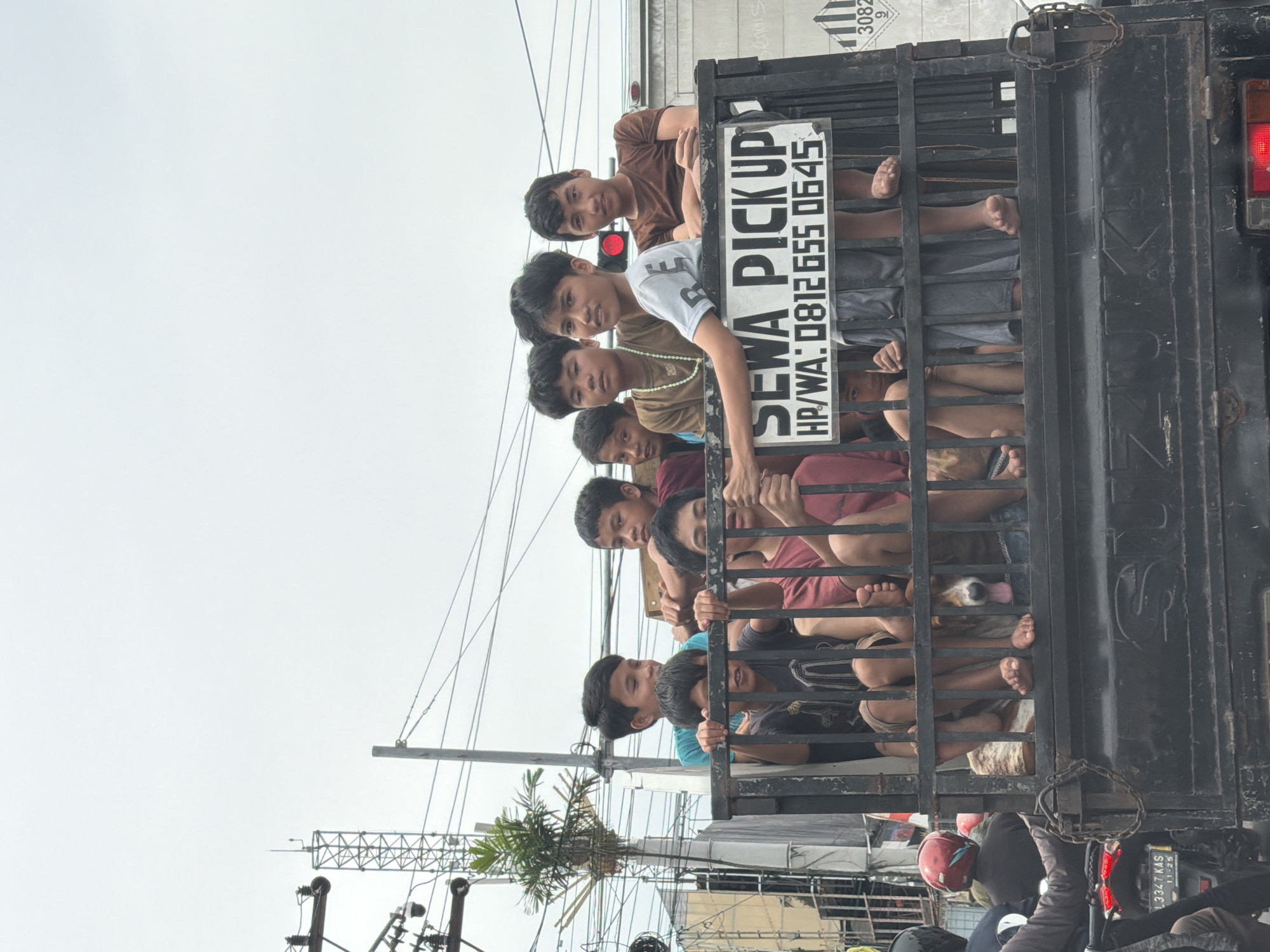 This media file depicts a group of young people, mostly boys, crowded into the back of a truck. The truck appears to be an open-bed pickup truck with a cage-like structure at the back. The individuals are seen peering over the railings and standing within the confines of the truck. Some are casually dressed in t-shirts and shorts, while others are in tank tops. Their expressions are varied, with some looking curious and others more stoic. The lighting suggests it is daytime, possibly overcast given the bright, diffused light. In the background, there are glimpses of urban elements, including overhead wires, poles, and possibly some buildings, indicating an outdoor, possibly street-side setting. A sign on the back of the truck reads "SEWA PICKUP HP/WA: 0812655 0645". This suggests the truck is available for rent. There is also a license plate visible, reading "13347 KAS 11-25". The overall impression is one of a group being transported, possibly for an outing or a group activity, with a sense of shared experience among them.