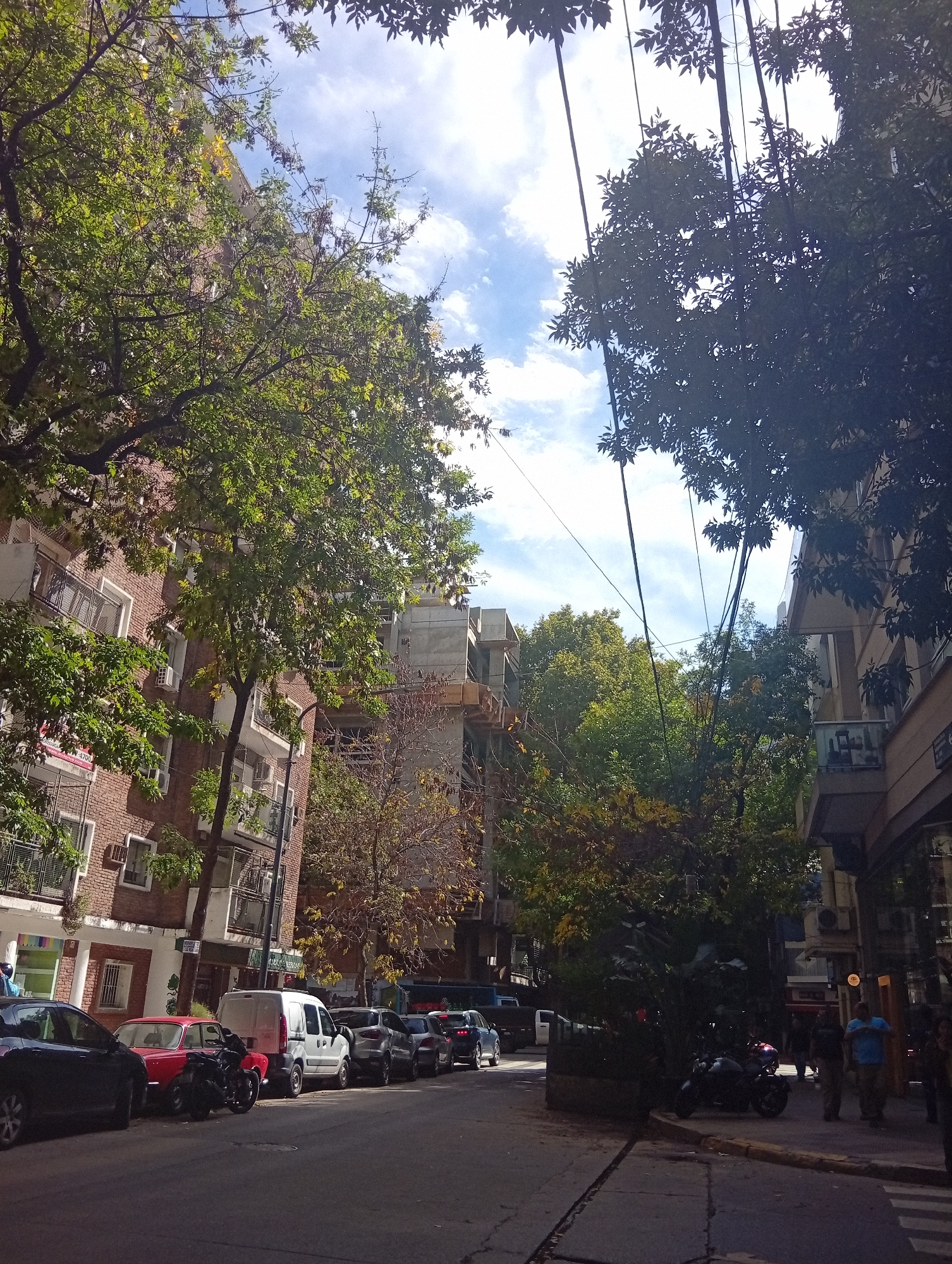 The image shows a street in Buenos Aires, Argentina. On the left is a brick building with balconies and awnings, partially obscured by green trees. A variety of cars are parked along the curb, including a red car and a white van. In the center, a building under construction is visible through the trees. On the right side of the street, a darker building is present, with people standing on the sidewalk. A motorcycle is parked on the sidewalk. Overhead, electrical wires stretch across the sky, which is visible through the gaps in the trees and shows some clouds. The street appears to be paved and has a curb separating it from the sidewalk. The lighting suggests it is daytime, with shadows cast by the buildings and trees.