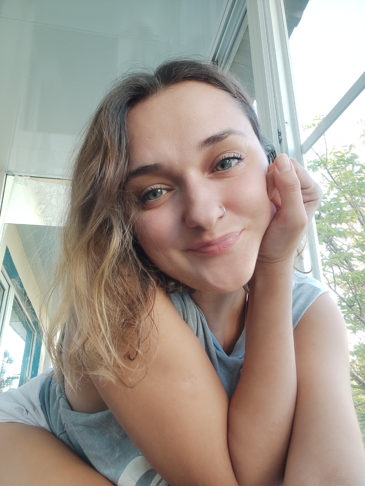 A young woman is posing for a photo indoors. She has light brown hair, light skin, and green eyes. She is wearing a light blue shirt and has a silver nose ring. Her left hand is supporting her head. The background shows a window with a view of a tree with green leaves. The location is not discernible as being in Yekaterinburg, Russia.