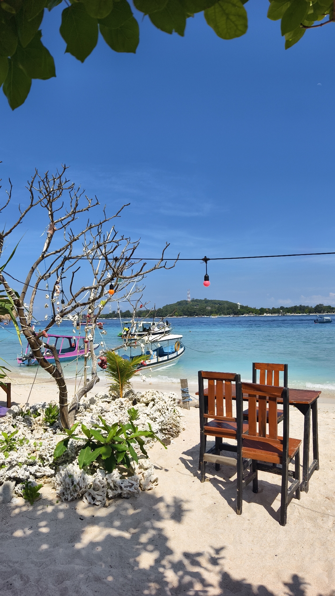A daytime shot on a beach in Unknown, Indonesia, shows a table and chairs at the edge of the beach with the ocean in the background. The sky is clear and blue, and the water is a light turquoise color. Several boats are visible in the distance. In the foreground, there is a bare tree with decorations hanging from its branches. There are also plants and coral visible in the sand. The table and chairs are made of wood and metal, and they cast shadows on the sand. A string of red lights are strung above the table.