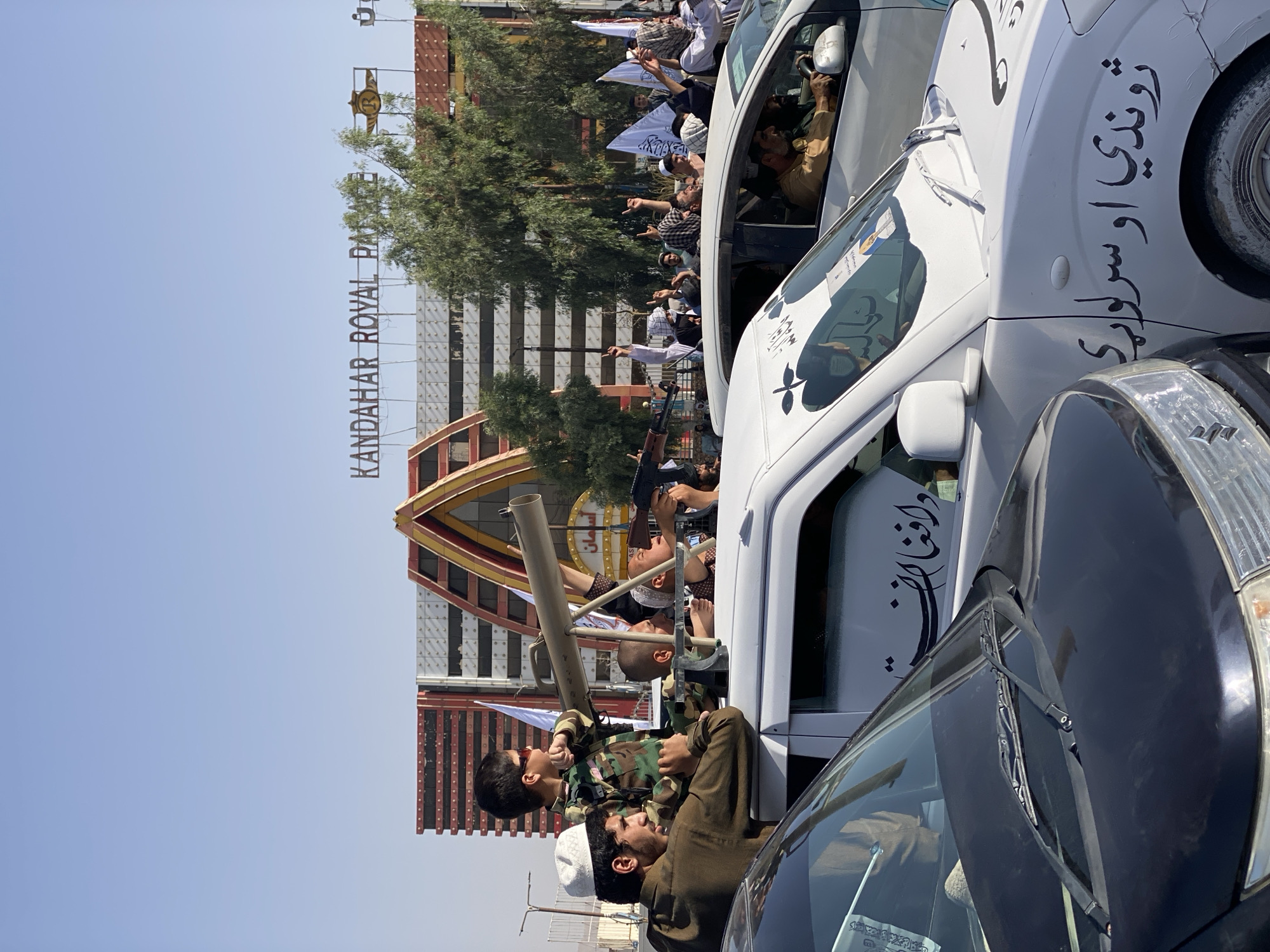 The image depicts a street scene in Kandahar, Afghanistan, with a group of people on top of and inside vehicles. In the foreground, a dark-colored car is partially visible. Behind it, a white car has several individuals. A young boy wearing a military-style uniform and sunglasses is standing on the roof of a white vehicle, holding what appears to be a rocket-propelled grenade launcher. Another man in a brown shirt and white cap is beside him. In the background, a building with the text "KANDAHAR ROYAL PALACE" is visible. Several white flags, some with inscriptions, are being held up by people in the crowd. Some individuals are gesturing with their fingers pointing upwards. The environment appears to be a sunny day with clear skies.