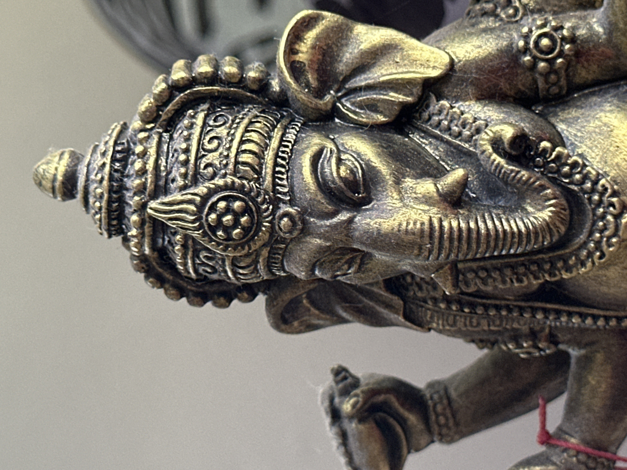 A close-up, waist-up shot shows a bronze statue of the Hindu deity Ganesha, characterized by an elephant head and human torso. The statue is intricately detailed with ornate jewelry and a crown. The background is a plain, light-colored wall. There are no people or animals present in the image, and the scene appears to be indoors, possibly on a shelf or altar. The statue is depicted from the chest up, with its trunk and one hand prominently visible. The statue is positioned slightly to the right of the frame's center. The lighting highlights the metallic sheen of the bronze. There is no indication of the city of Karon, Thailand within the image itself.
