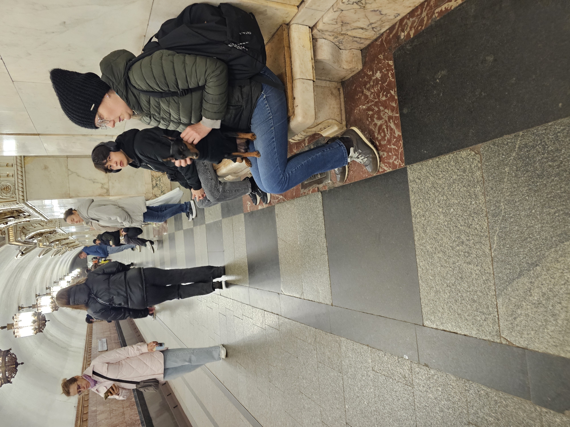 A vibrant scene unfolds within what appears to be a grand metro station, likely in Moscow given the provided context. The image, originally rotated, shows several individuals and a small dog on a tiled platform.
In the foreground, a person wearing a black knitted beanie, glasses, and a dark green puffer jacket sits or leans on an ornate marble bench or ledge. Beside them, another person in a black hoodie and headphones is also seated or leaning, holding and petting a small, black dog with tan markings on its legs and chest. A black backpack rests nearby. The marble wall behind them is white with reddish-brown veining, transitioning to an older, light brown, decorative section at the base.
Further down the platform, several other people are visible. One person in a light grey jacket and blue jeans stands, while another in a black puffer jacket and black pants waits further along. In the mid-ground left, a person wearing a light pink quilted jacket, light blue jeans, and glasses looks at their phone. More indistinct figures can be seen in the distance along the arched tunnel.
The station's architecture is notable, featuring ornate chandeliers hanging from the high, arched ceiling, decorative molding, and columns, all indicative of a historically rich and grand public space. The floor is covered with various types of stone tiles in shades of grey, black, and reddish-brown. The overall atmosphere suggests people waiting casually in a beautifully designed, busy transit hub. No significant readable text is visible, although some faint, unidentifiable text appears on one person's jacket.