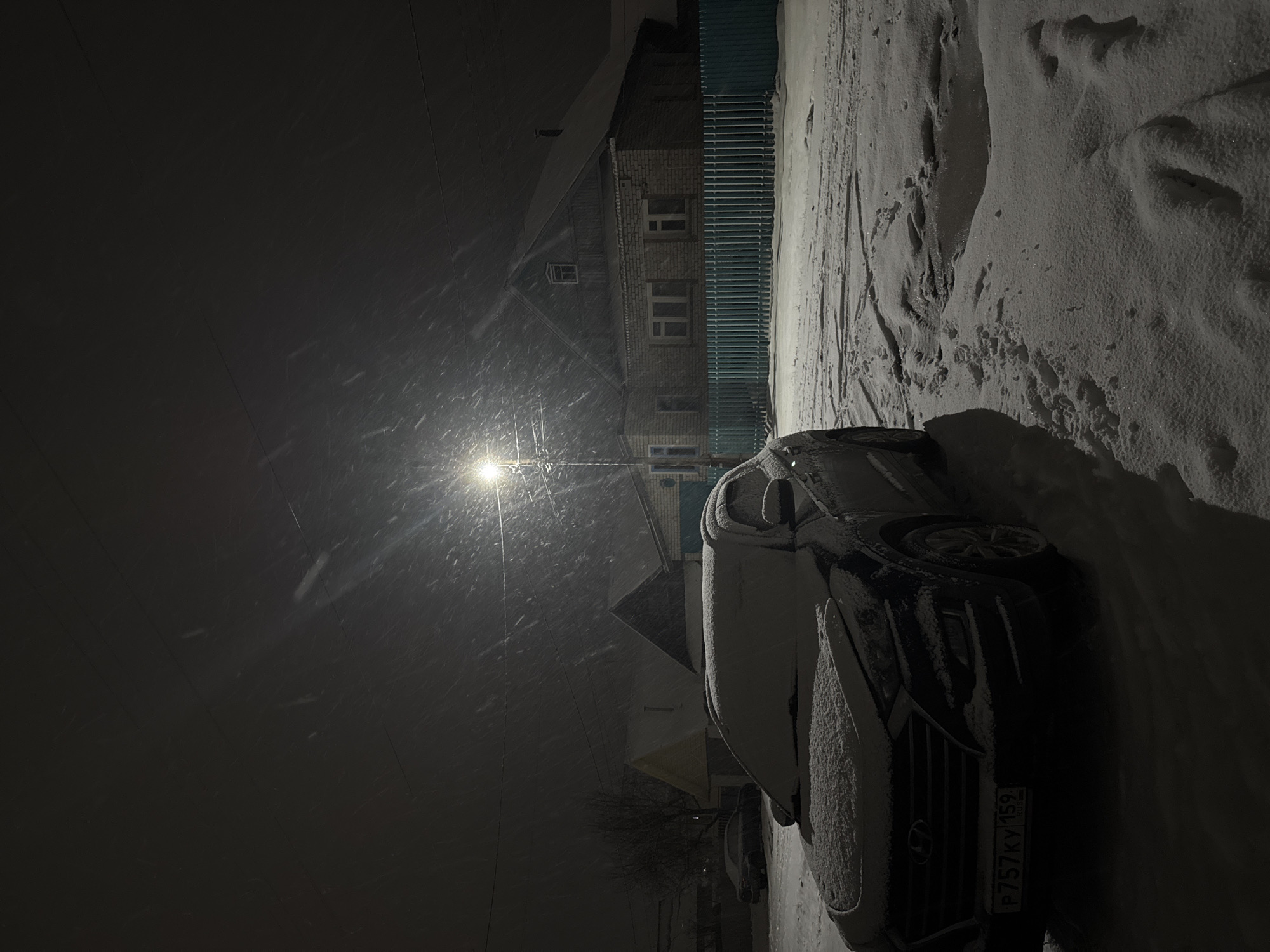 The image shows a snowy street scene at night in the city of Chaikovsky, Russia. A car, partially covered in snow, is parked on the side of the road. In the background, there is a brick building with a fence and some trees. A street lamp illuminates the falling snow, creating a bright spot in the otherwise dark scene. The license plate on the car reads "P757 KY 159 RUS". Footprints are visible in the snow.