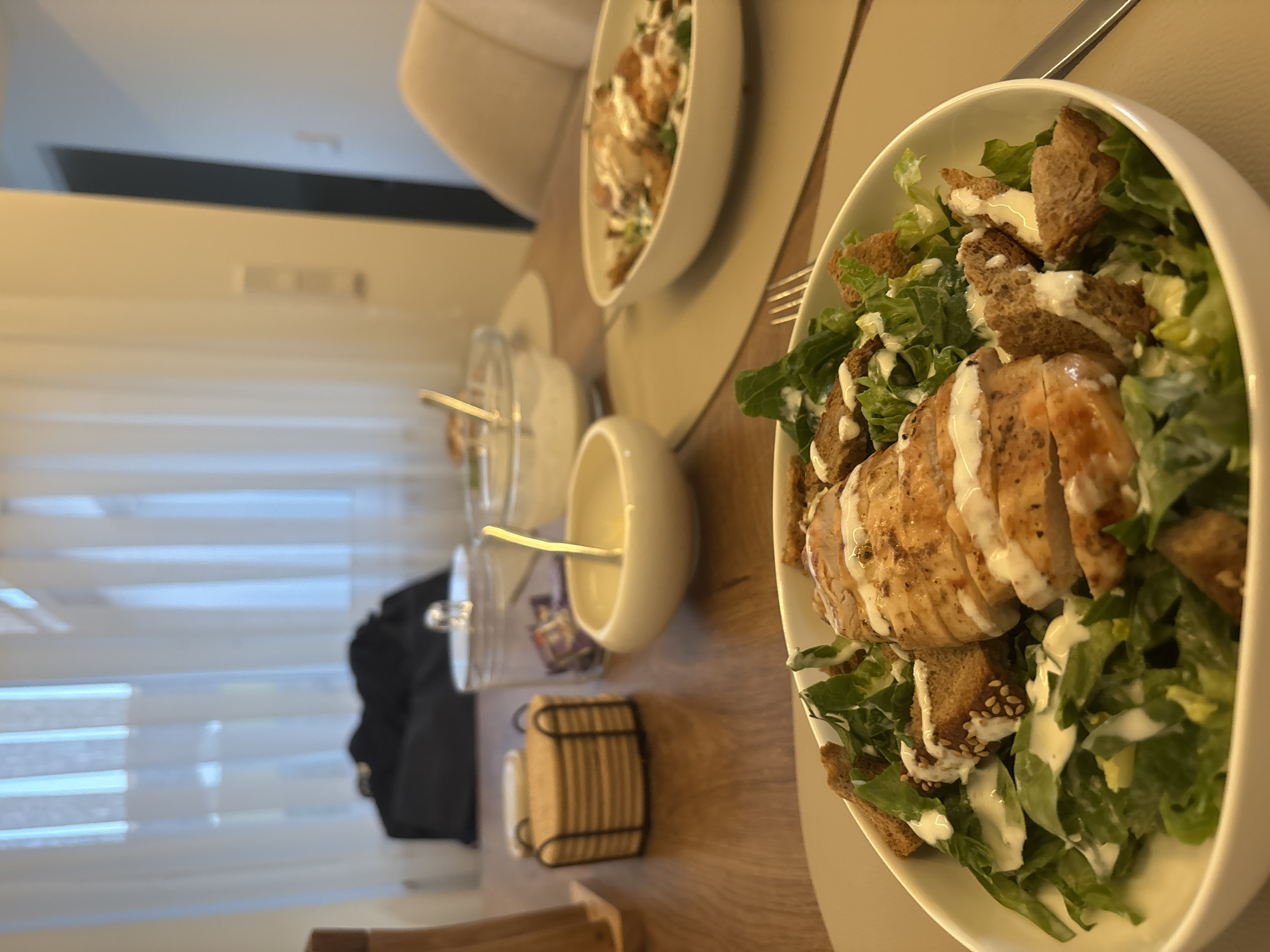 A close-up shot shows two bowls of Caesar salad on a wooden table. The salad in the foreground is in a white bowl and contains sliced grilled chicken, romaine lettuce, croutons, and a creamy dressing. The second bowl, further back, is also white and contains a similar salad. In the background, a stack of coasters and a glass jar with a lid are visible. A pair of chairs and a curtained window are also present in the scene. The setting appears to be a dining area in a residential building.