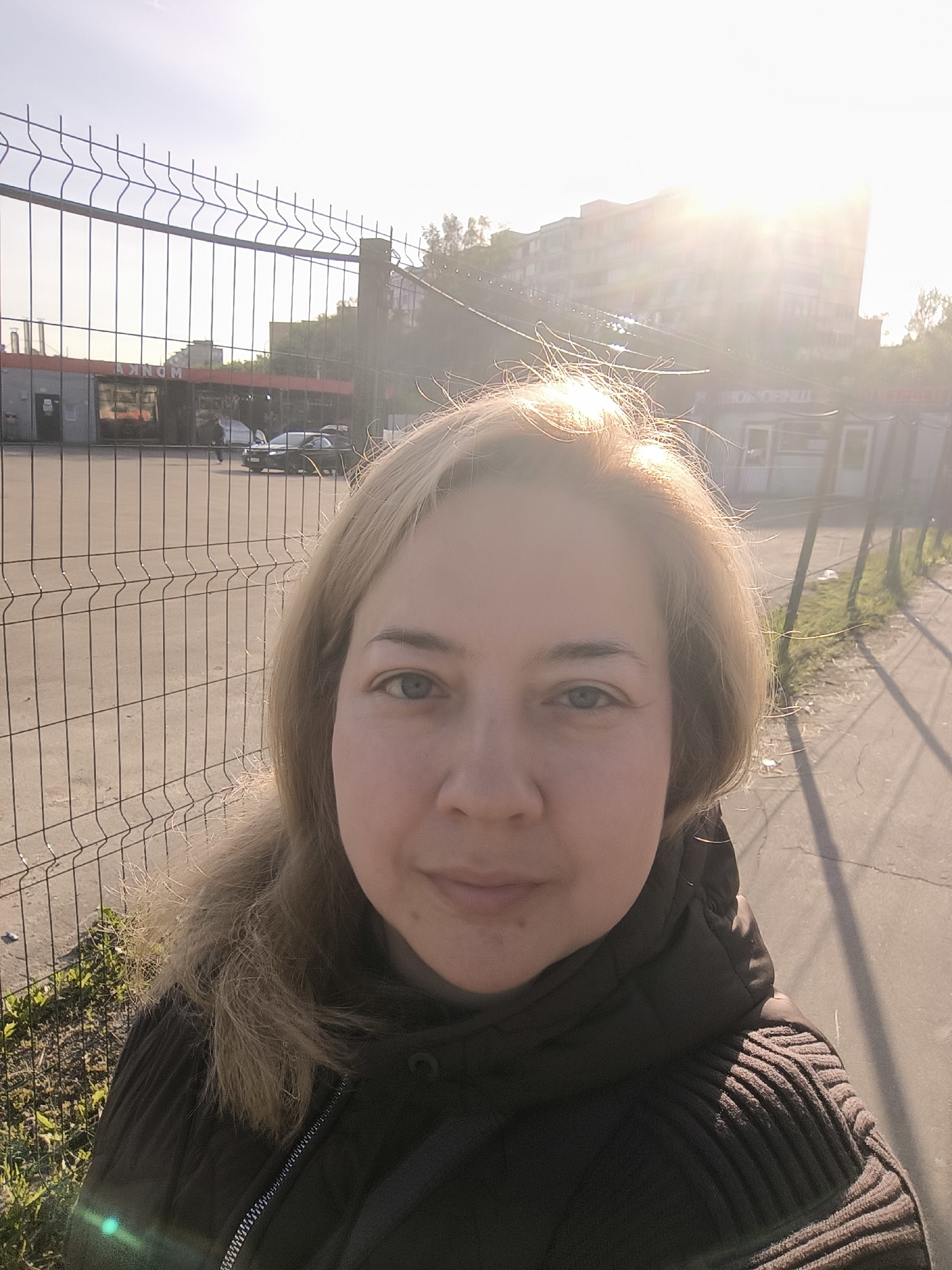 A woman stands in the city of Unknown, Unknown. She is positioned beside a chain-link fence, wearing a dark hooded jacket. The sun is visible in the background, positioned behind buildings and causing glare. The area appears to be a street or path, with buildings, parked vehicles, and some greenery visible through the fence.