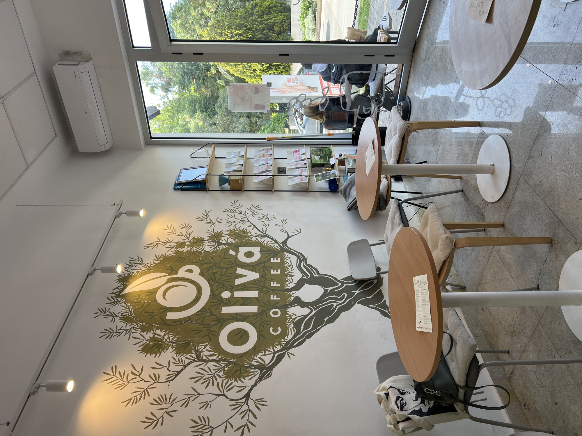 The photograph shows the interior of a cafe in Bar, Montenegro. The cafe's wall features a stylized tree mural incorporating the words "Oliva Coffee". The space is bright with natural light from a large window. Several small round tables with accompanying chairs are arranged on a light grey tiled floor. A shelf unit is visible on a wall near the window. Through the window, a street scene with vegetation and parked vehicles can be seen. An air conditioning unit is mounted high on the wall.
