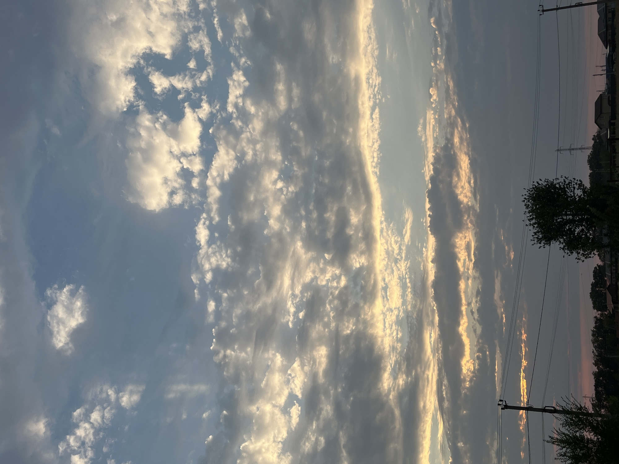 The image depicts a cloudy sky above a rural landscape. The clouds are varied, with some appearing white and illuminated by sunlight, while others are darker and more ominous. The sky is a mix of blues and grays. Below the sky, a line of trees and houses is visible, suggesting a residential area on the outskirts of a city. Electrical poles and wires run across the scene, indicating the presence of infrastructure. The overall impression is one of a natural landscape with signs of human habitation. The specific location within Taganrog City District, Russia is not discernible from the image alone.