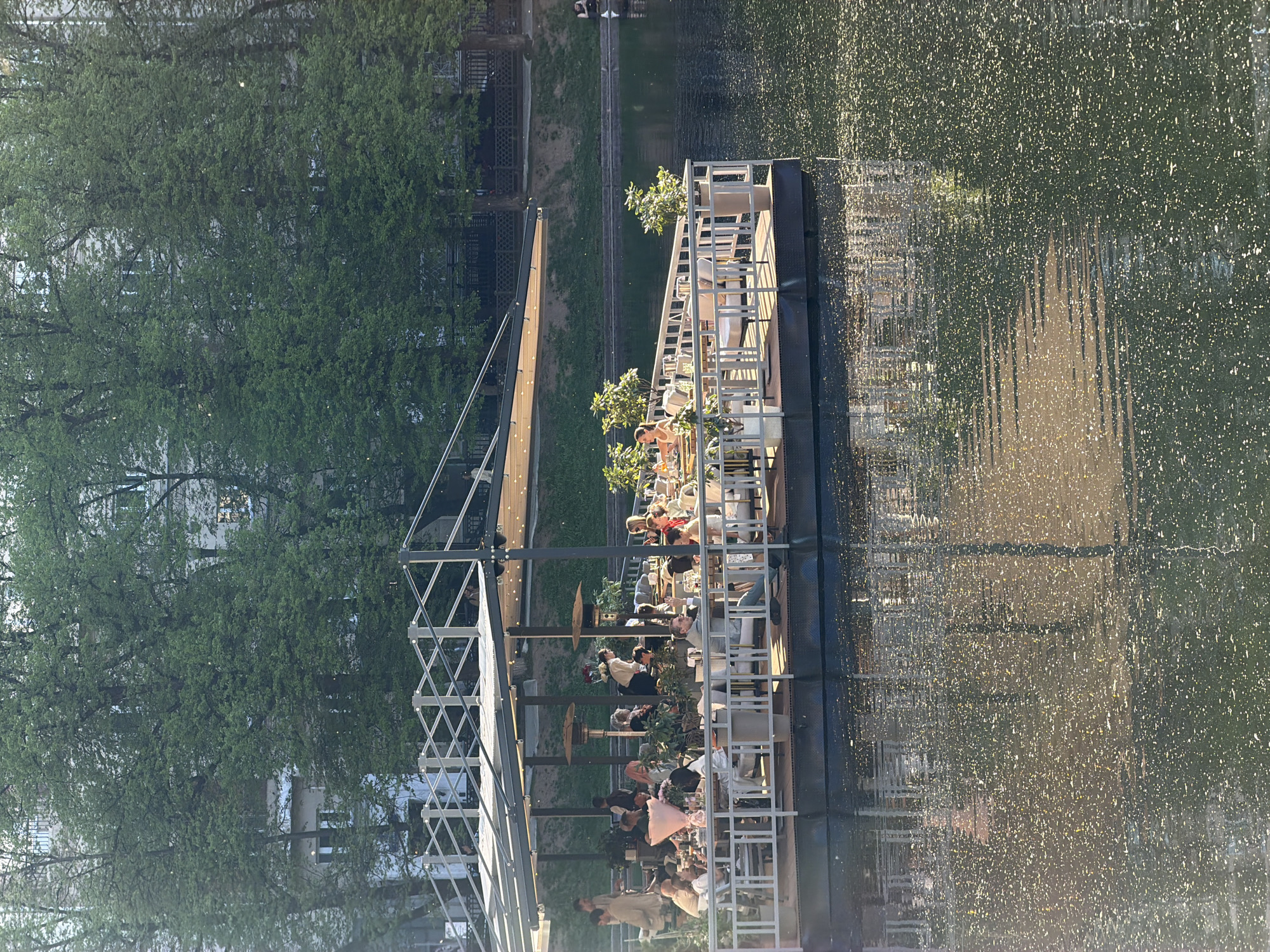 The image depicts a floating restaurant on a body of water, likely a canal or the Moscow River, in Moscow, Russia. The restaurant is constructed on a barge-like platform with a white frame structure supporting a roof. People are seated at tables, engaged in conversation and dining. Trees line the bank adjacent to the floating structure. The water reflects the surrounding environment, showing the sunlit surface and the structures of the restaurant.