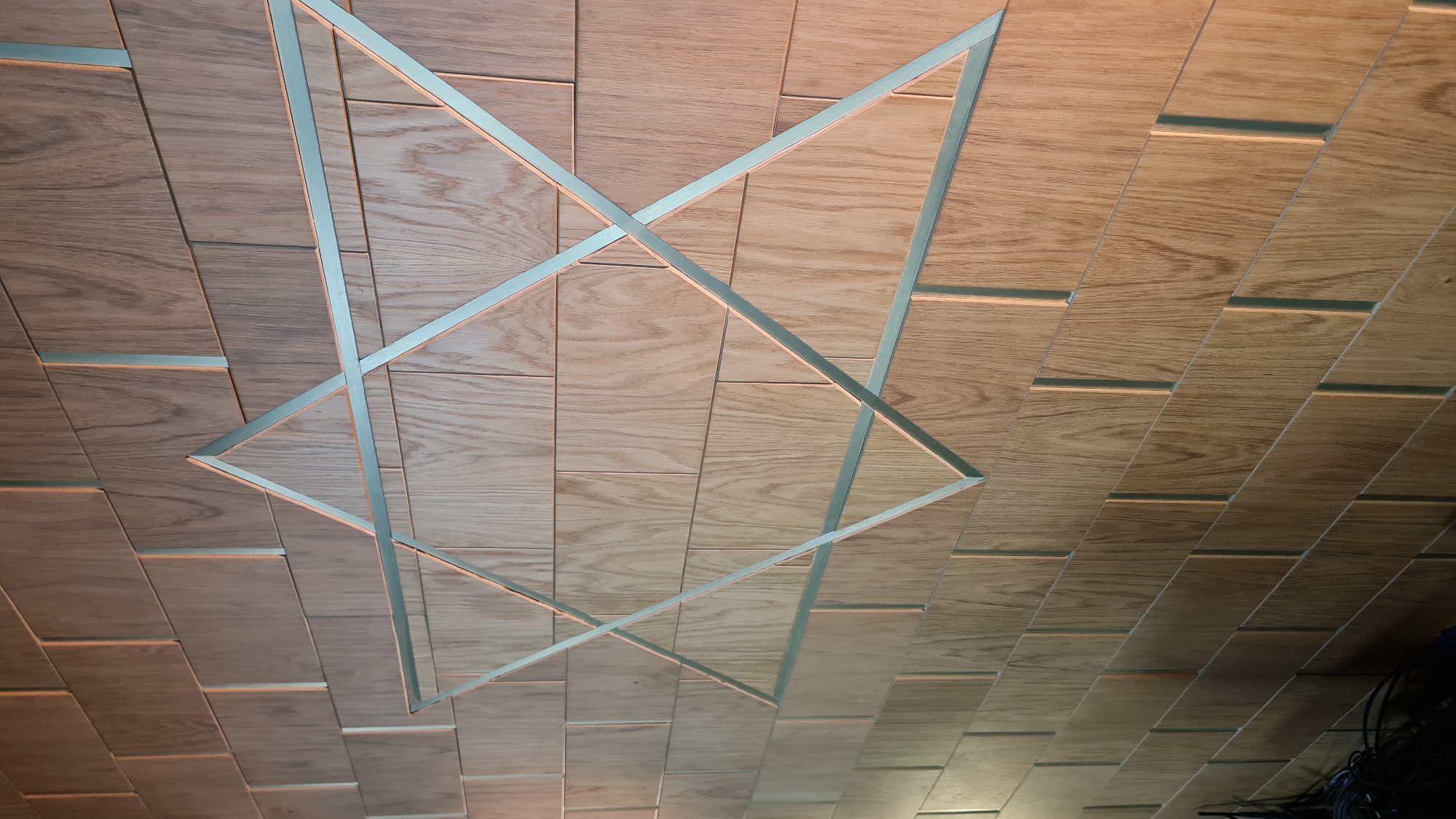 A low-angle, indoor shot depicts a ceiling covered with rectangular wooden tiles interspersed with narrow strips of silver-colored material. A geometric structure made of similar silver material is suspended from the ceiling. Cables are visible in the lower right corner. No people are visible. There is no information to indicate a location in Nahariyya, Israel.