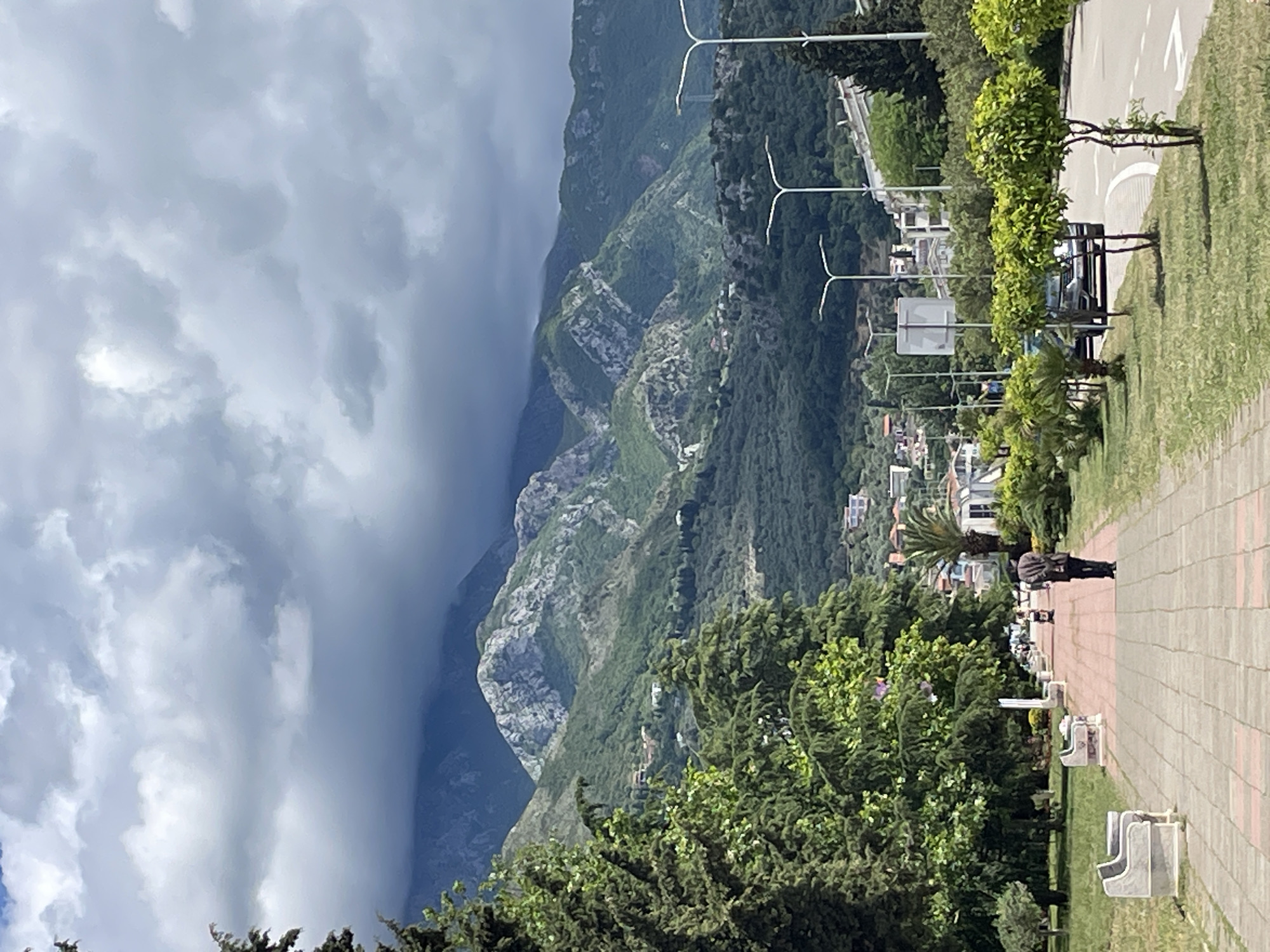 The image depicts a scenic view in the city of Bar, Montenegro. A brick pathway stretches from the bottom right towards the middle ground, lined with benches and manicured greenery to the left. A lone individual is walking on the path in the middle ground, away from the camera. Beyond the pathway, a cityscape of white and light-colored buildings is visible. Mountains rise dramatically in the background, partially obscured by heavy, dark clouds filling the sky. A row of light poles follows a road in the mid-ground. The overall atmosphere suggests a cloudy day in a mountainous, coastal region.
