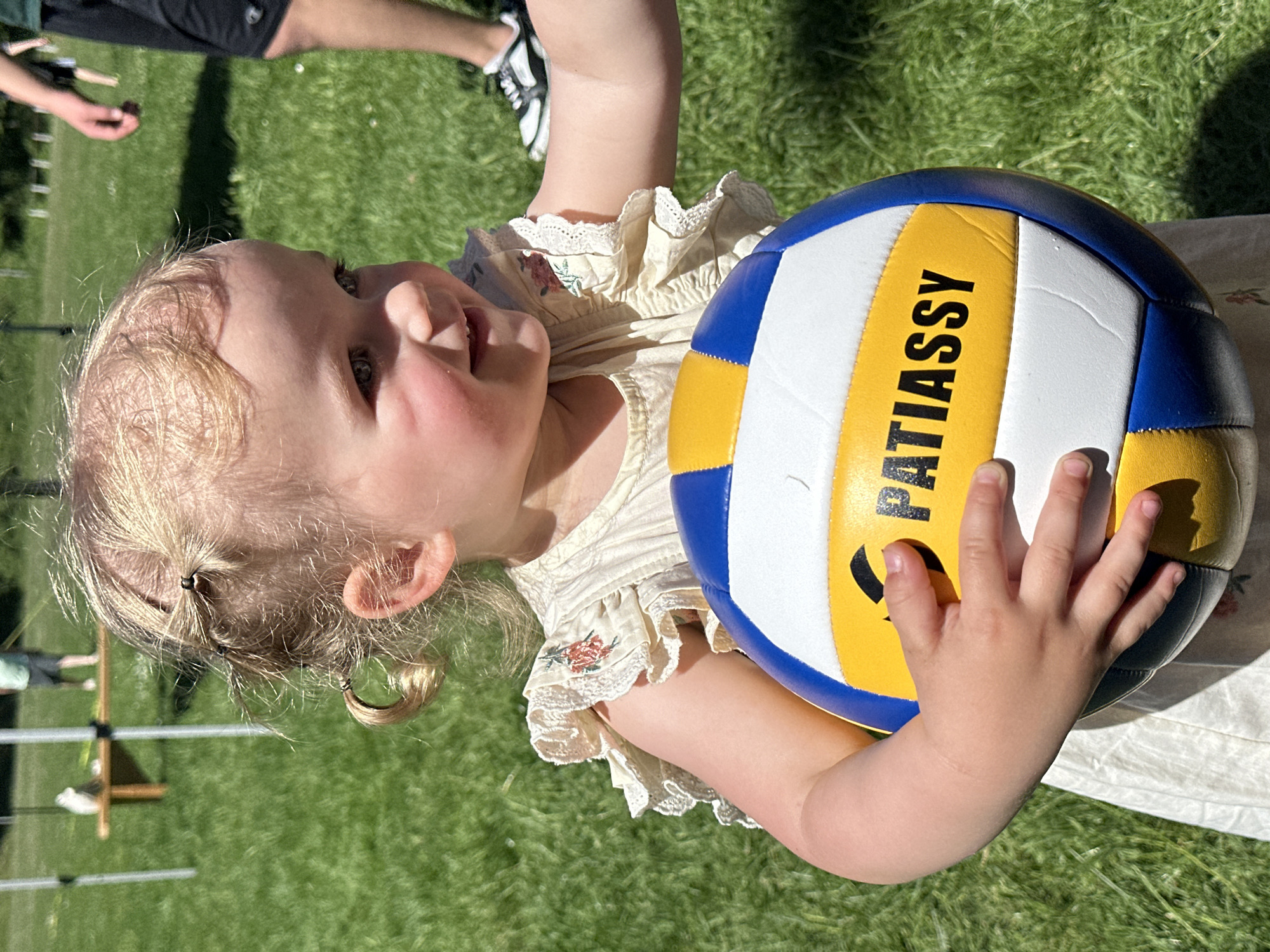 A toddler with blonde hair in pigtails is holding a blue, white, and yellow volleyball that reads "PATIASSY" in black capital letters. The toddler is wearing a light-colored dress with lace trim and small floral details. They are positioned on a green grassy surface, likely a lawn. The scene appears to be outdoors on a sunny day. Part of another person's leg and foot wearing black and white sneakers is visible in the background. Additionally, a partially visible structure resembling a hurdle or small fence is located in the background. Given the prompt, the location is specified as Grünberg, Germany.