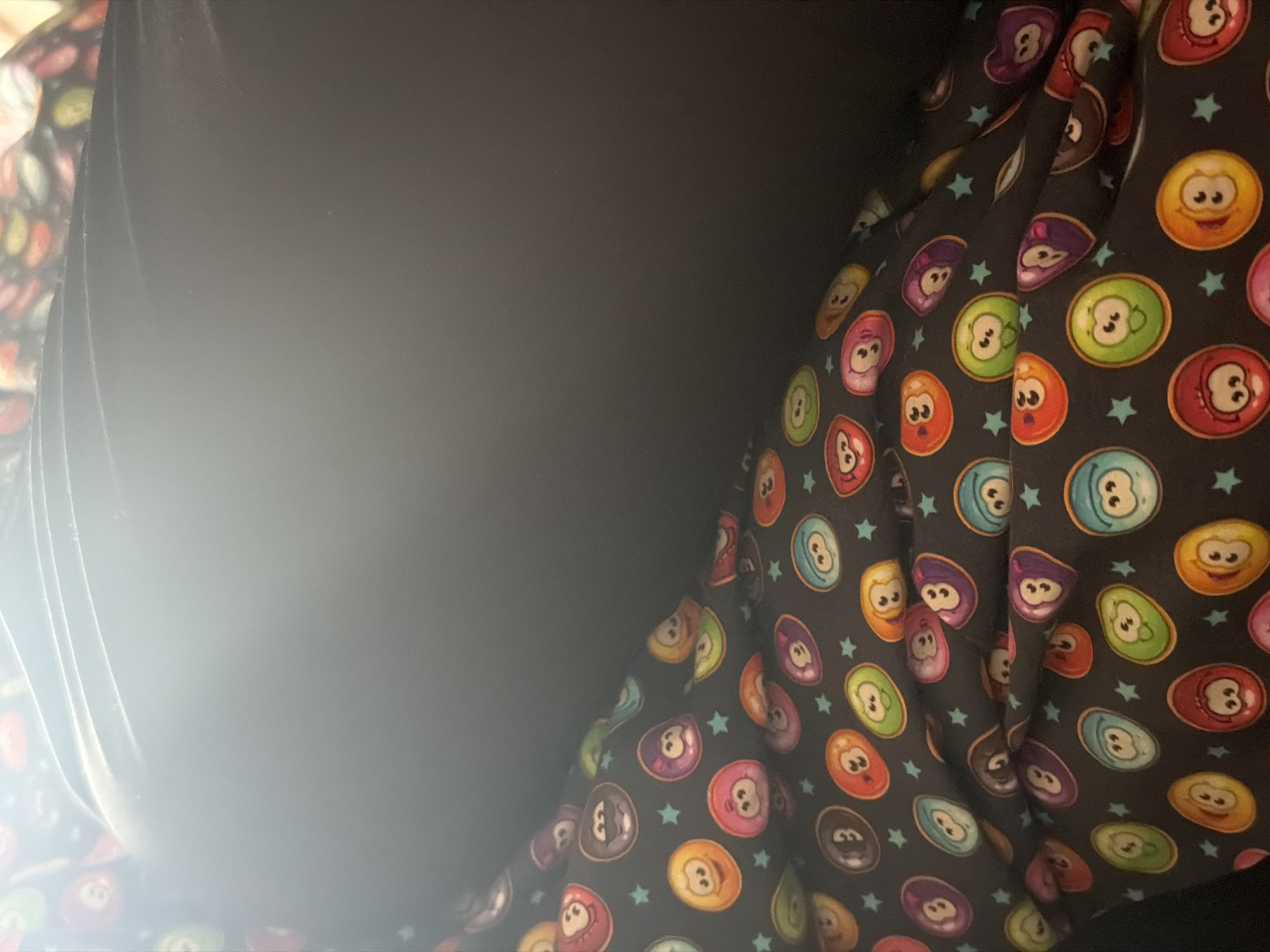 The image presents a close-up view primarily featuring a dark fabric patterned with an array of colorful cartoon characters and small stars, alongside a large, dark, indistinct area. The patterned fabric is visible on the right and bottom-left portions of the frame, exhibiting folds and creases. The cartoon characters are oval or slightly bean-shaped, resembling stylized pool balls or fantasy creatures, depicted in various vibrant colors such as red, green, orange, purple, blue, yellow, and pink. Each character features distinct white eyes and a small mouth, with some displaying additional small circular markings or "ears." Scattered across the dark background of the fabric are small, teal-colored star shapes.
The central and left areas of the image are dominated by a dark, featureless expanse, possibly a plain wall or another piece of fabric. A bright, overexposed area is present on the far left, suggesting a strong light source or reflection, partially obscuring a dark, folded material. The scene is static, focusing on an object rather than an activity or event. There are no clear indicators within the image itself regarding the time of day, weather conditions, or specific location cues beyond the provided context of Iskar, Bulgaria. The expressions on the cartoon characters are generally cheerful or neutral. No visible text is present in the media file.