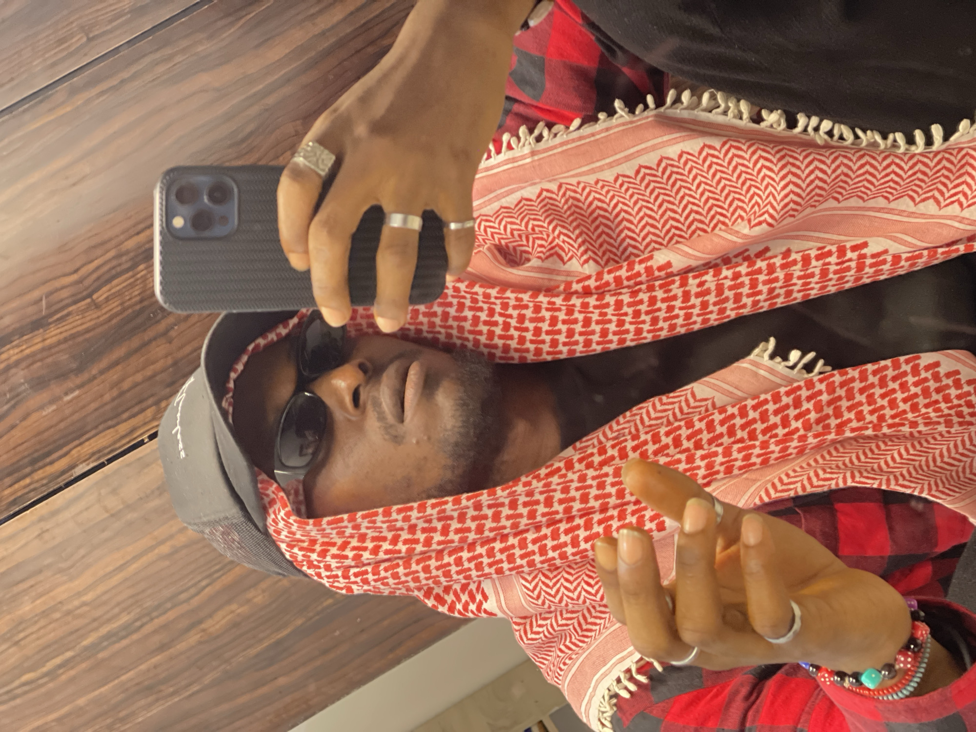 A selfie is taken by a Black man in a room with a wooden paneled wall. He is wearing a black baseball cap, sunglasses, and a red and black checkered shirt over a black t-shirt. A red and white checkered keffiyeh is draped around his head and neck. He is holding a black smartphone in his right hand, taking the picture. He is wearing several silver rings on his fingers. His left hand is raised, with fingers extended. There are beaded bracelets on his left wrist. The overall impression is one of casual style. The location is identified as Abuja, Nigeria.