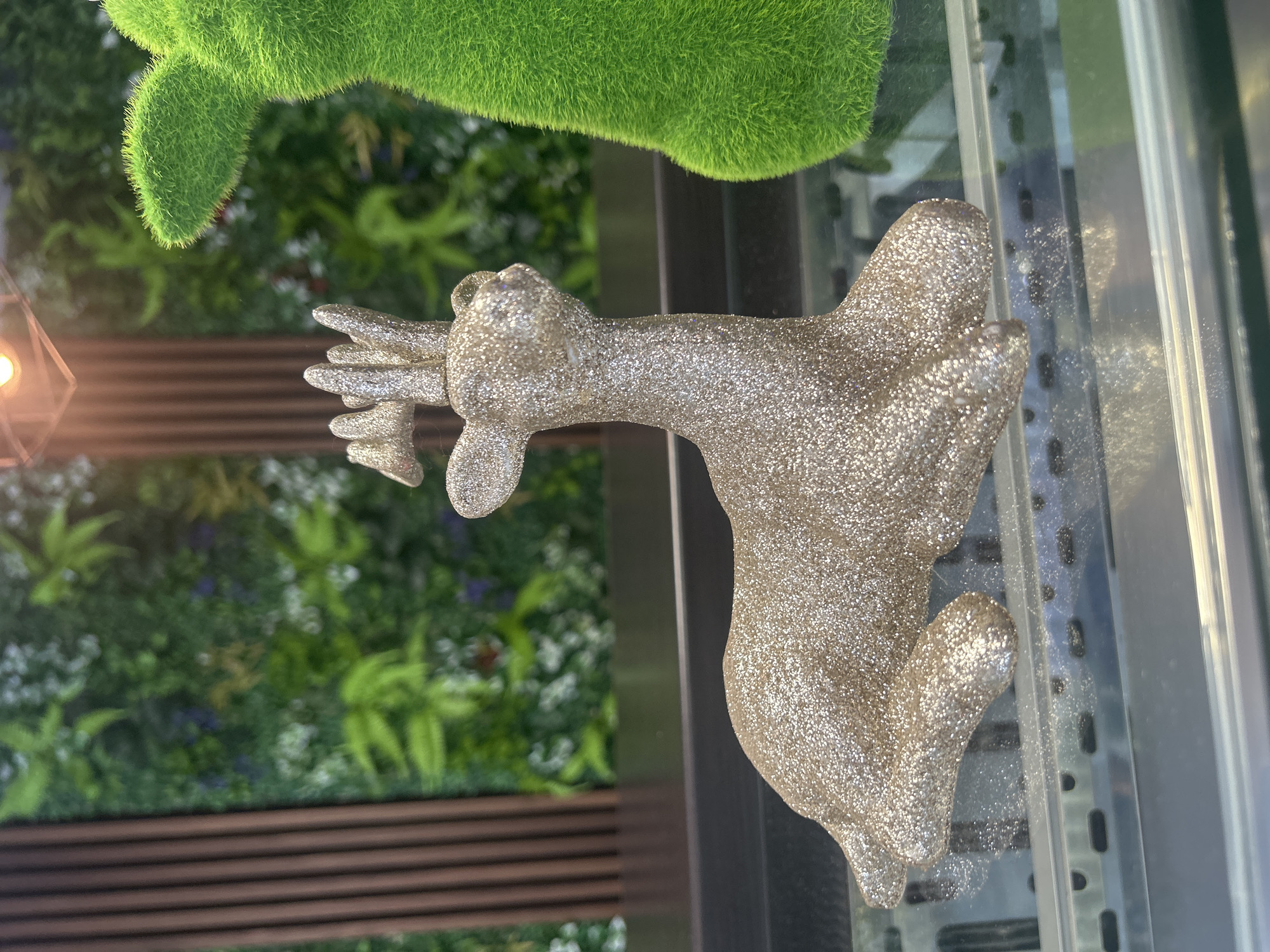 A glitter-covered, gold-colored reindeer figurine is positioned on a glass shelf in a display case. The reindeer is lying down, facing left, with its head slightly raised and turned towards the viewer. Behind the reindeer, a fuzzy green object, possibly another figurine or a decorative plant, is partially visible on the right side of the frame. The background consists of a blurred arrangement of greenery and wooden vertical elements, suggesting an indoor setting with decorative plants. The lighting appears to be artificial, casting a warm glow. The image does not provide any specific information about its location in Varna, Bulgaria.