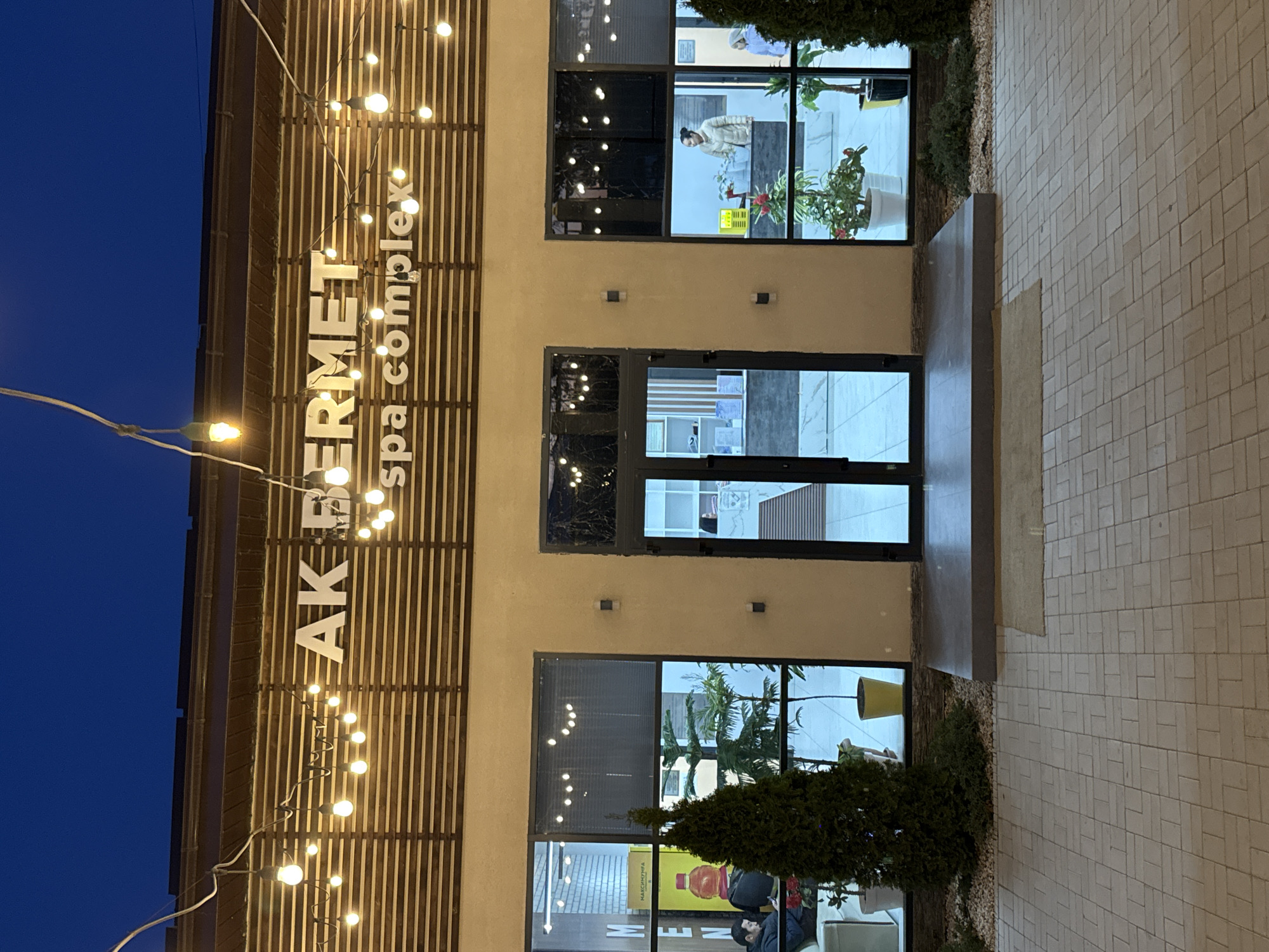 Here is a description of the image:
The image shows the exterior of a building in Kara-Oy, Kyrgyzstan, identified as "AK PERMET spa complex". The facade is beige with a vertical wooden slatted section on the left, illuminated by strings of light bulbs. Three sets of black-framed windows and a central doorway punctuate the facade. The doorway appears to lead directly inside with a small entryway area. The windows show interior details, including plants, furniture, and people. A man is visible in the bottom window. Greenery is planted at the base of the building, flanking the entrance. The ground is paved with light-colored rectangular stones. The sky visible in the upper left corner is a deep blue, suggesting it is either early morning or evening. The overall lighting of the scene is a mix of natural light and artificial light from the string lights. The camera angle is slightly tilted.