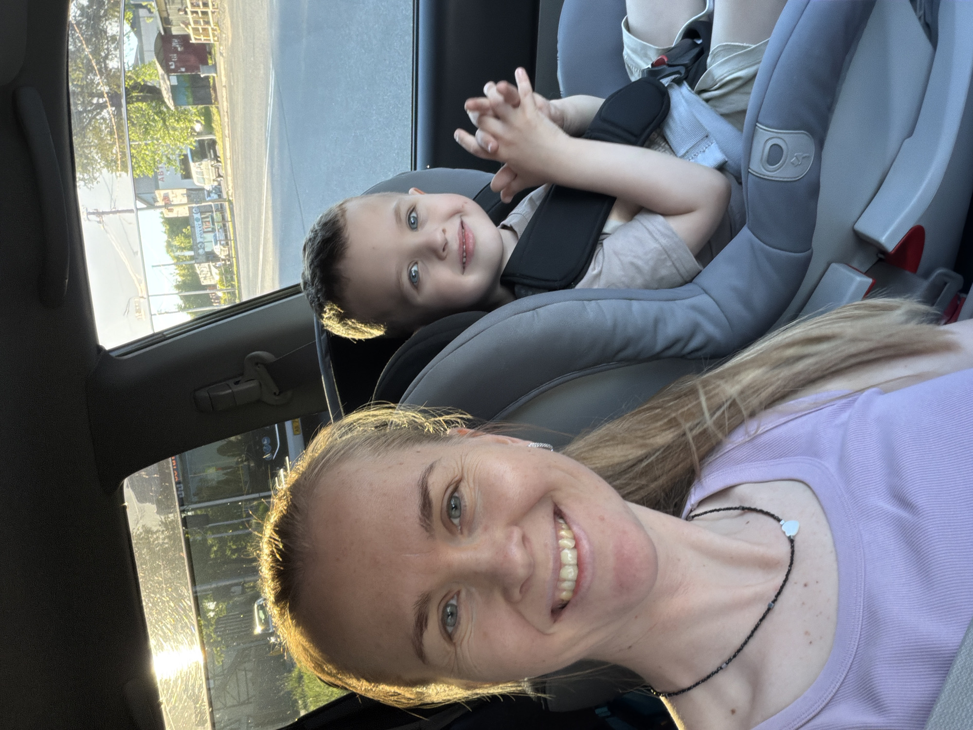 The media file shows a close-up, selfie-style shot taken from inside a car. In the foreground, a woman with long blonde hair smiles at the camera. She is wearing a light purple top and a black necklace with a small heart pendant. Behind her, a young boy with blue eyes is seated in a car seat. He is looking towards the camera and smiling, with his hands clasped in front of him. The boy is wearing a light-colored t-shirt. The setting appears to be daytime, with sunlight streaming in through the car window, illuminating the exterior scene. Outside the car, a street scene is visible, with buildings, trees, and a vehicle. The time of day is likely late afternoon or early evening due to the warm, golden light. The overall mood is happy and relaxed, suggesting a family outing. No visible text is present in the image. The context provided indicates the location is Khabarovsk, Russia.